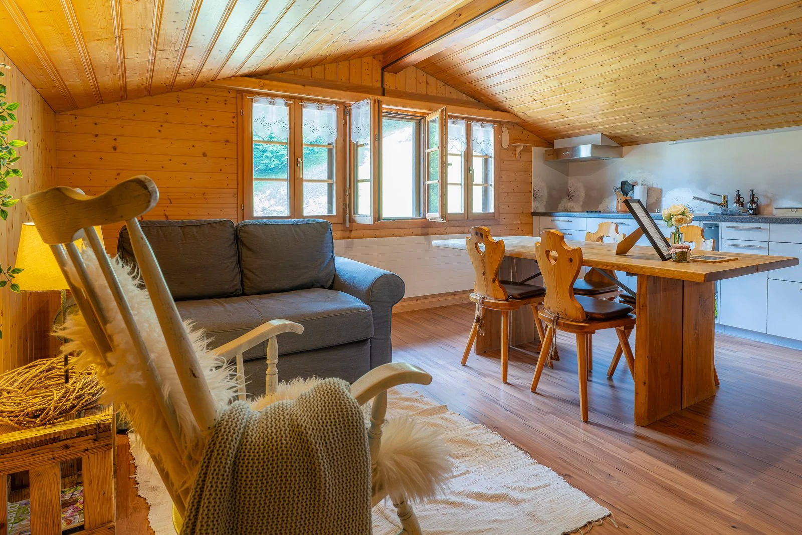 Gemütliches Wohnzimmer in einer Holzhütte mit einem grauen Sofa, Holzstühlen am Esstisch, offenen Küchenbereich, vielen Fenstern und warmer Beleuchtung.