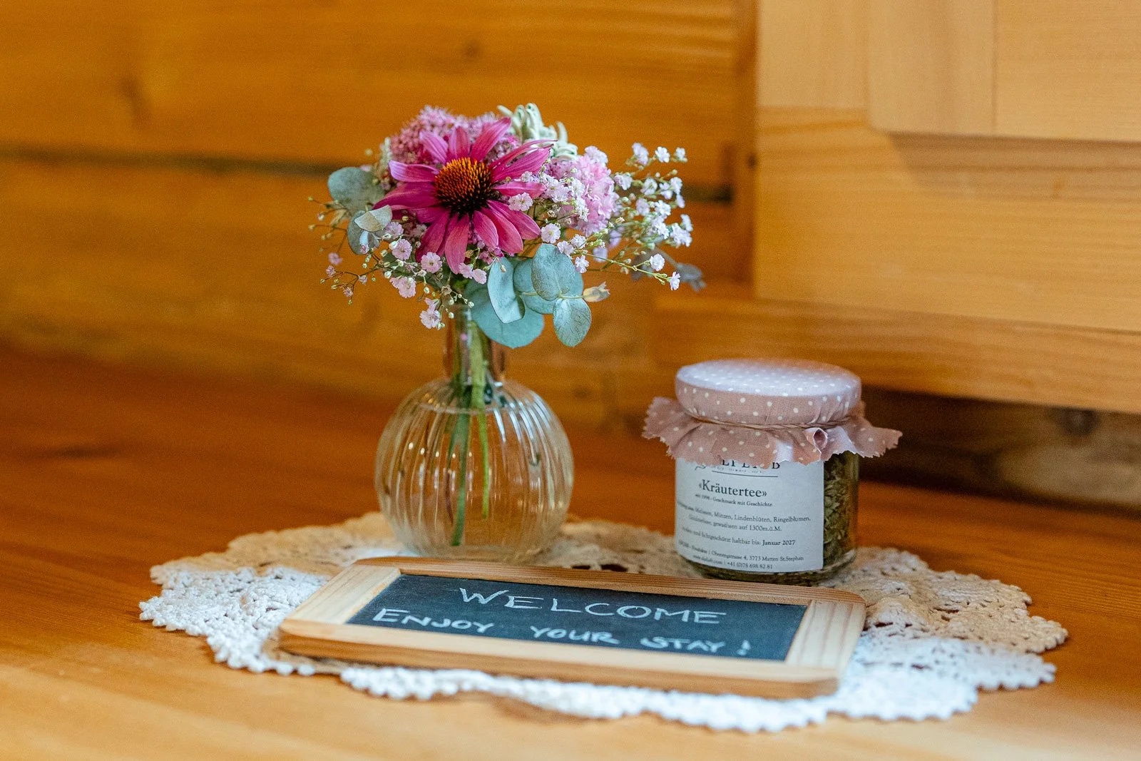Ein Blumenstrauß in einer Vase, ein Glasteller mit einer Flasche Kräutertee, eine kleine Tafel mit der Aufschrift "Willkommen, Genießen Sie Ihren Aufenthalt!" auf einer kleinen, handgefertigten Stoffdecke