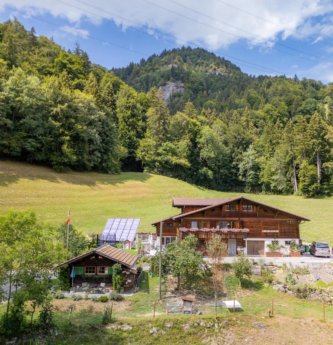 Ein Landhaus inmitten eines grünen Hügels, umgeben von Bäumen, mit Bergen im Hintergrund, blauer Himmel und Wolken.