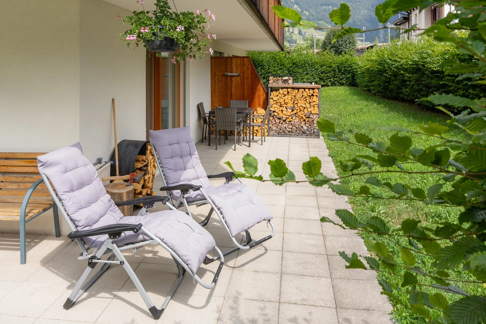 Blick auf eine Terrasse mit zwei bequemen Liegestühlen, einem Gartenstuhl und einem Gartentisch mit Stühlen. Im Hintergrund ist ein Holzstapel und ein großer, gepflegter Garten mit Bäumen und Büsche zu sehen, umgeben von einer grünen Landschaft.