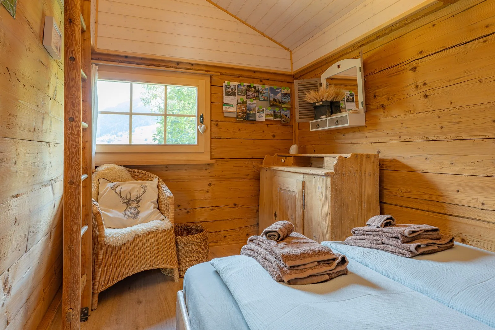 Ein rustikaler Schlafraum mit Holzverkleidung, einem Bett mit zusammengelegten Handtüchern, einem Sessel mit Kissen, einem kleinen Holzschrank und Dekorationen an der Wand, darunter ein Spiegel und eine Fotowand. Ein Fenster zeigt eine grüne Landscha