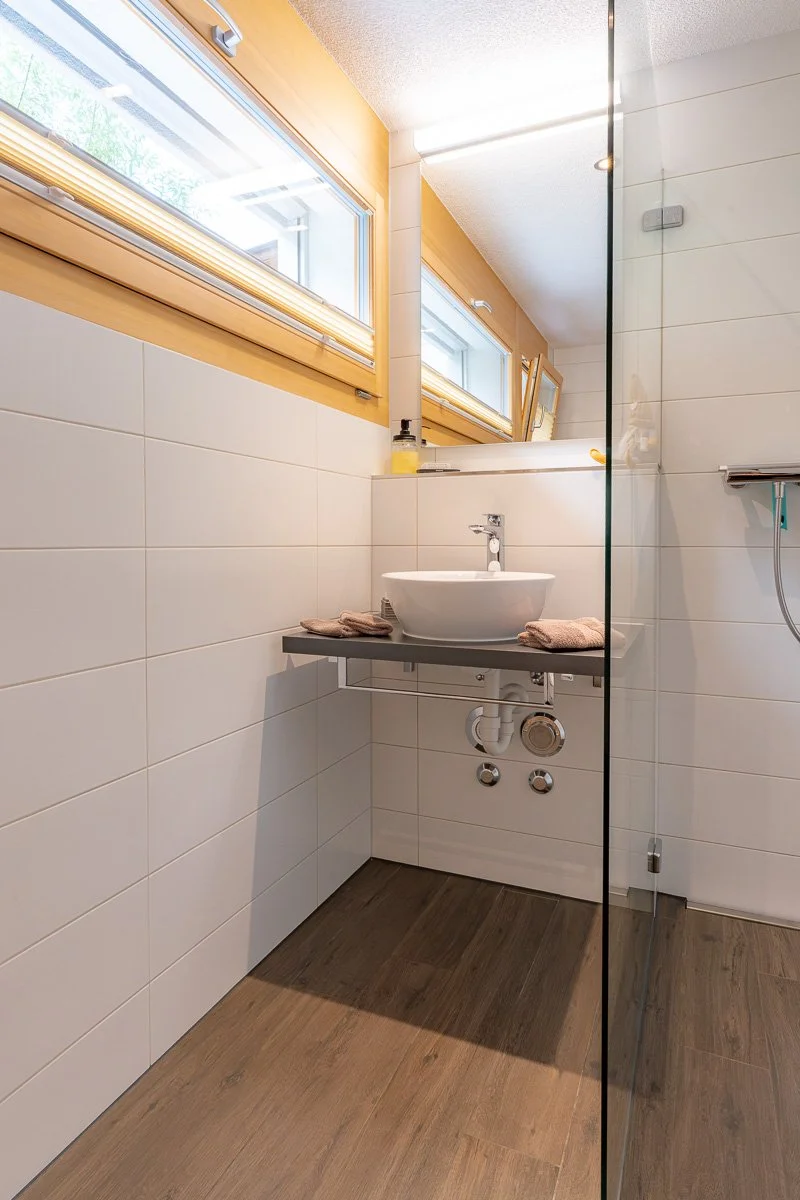 Ein Badezimmer mit weißen Wandfliesen, einem Fenster mit Holzkastenschalung, einem Waschbecken auf einem grauen Regal, einem Wasserhahn und Handtüchern.
