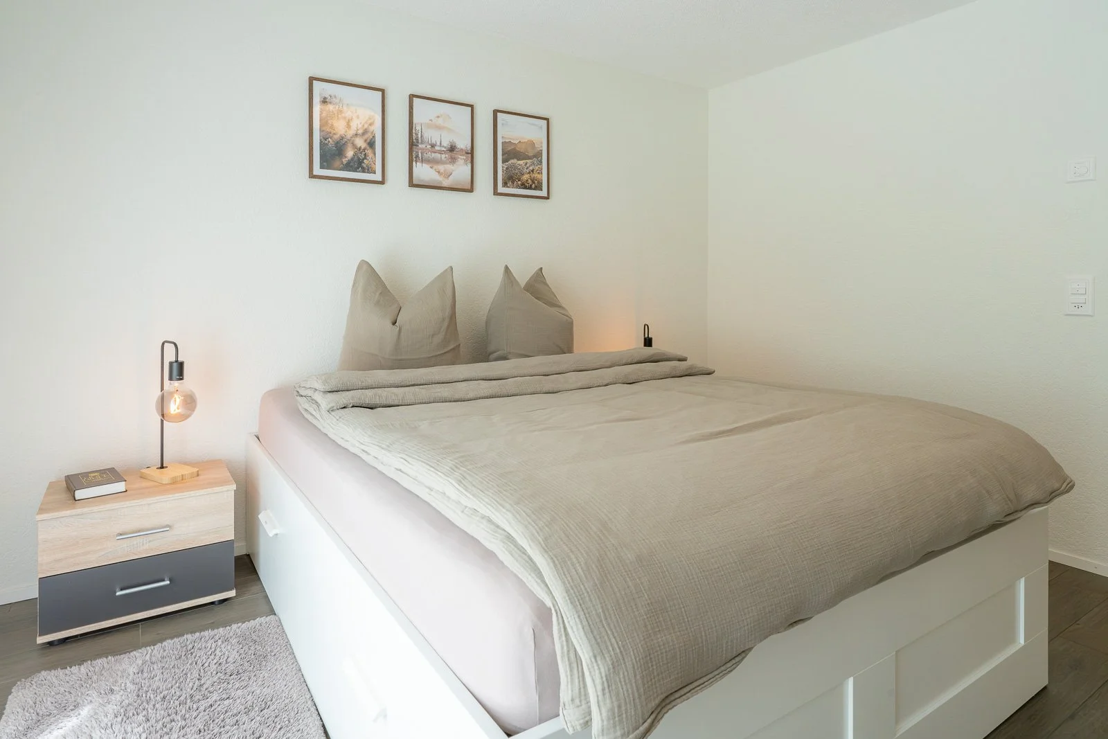Ein modernes Schlafzimmer mit einem großen Bett, zwei Kissen, Nachttisch mit Lampe, Buch und einem runden Teppich, weiße Wand mit drei Kunstwerken