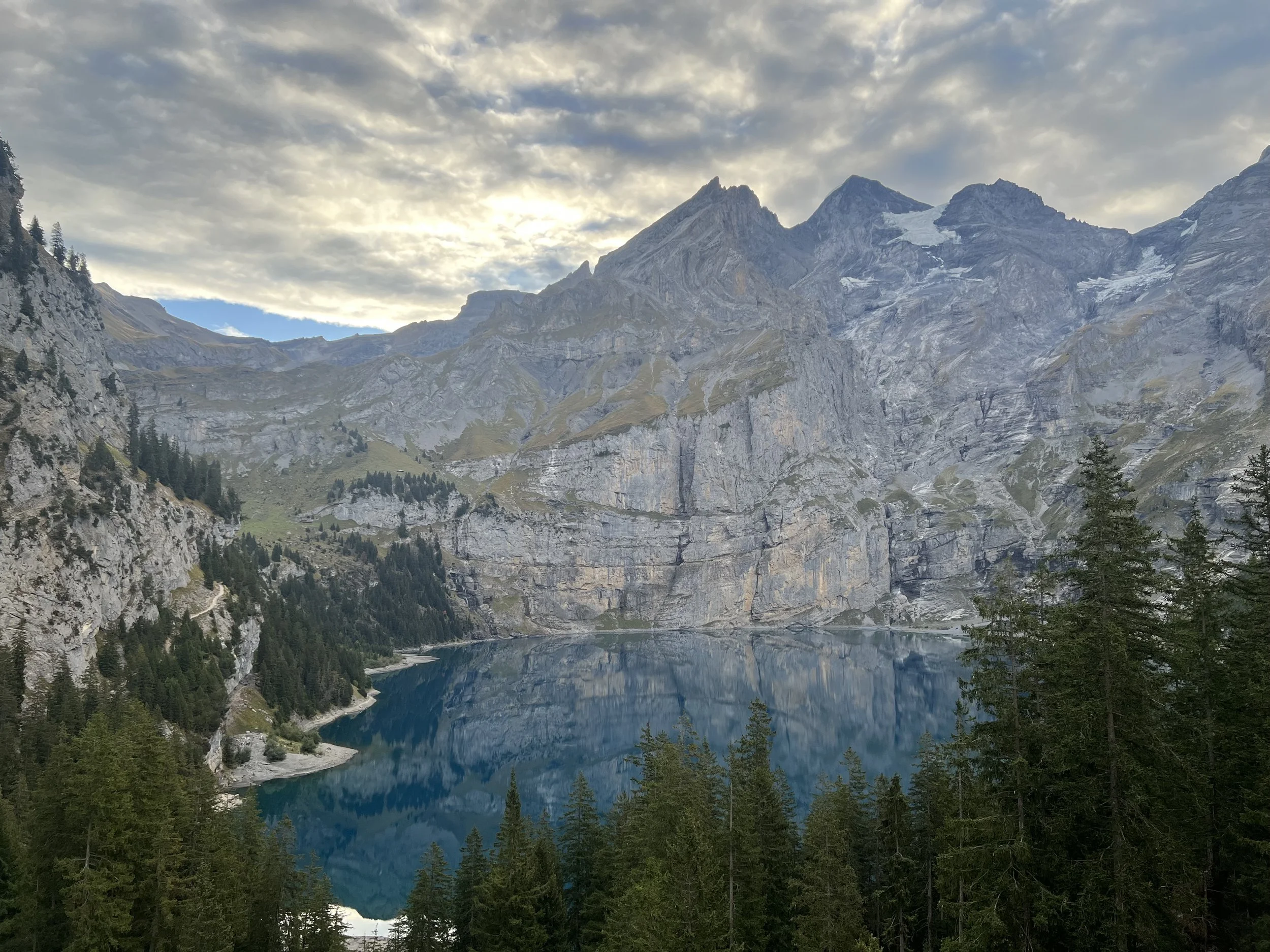 Bergsee mit Spiegelungen vor schroffen, teilweise vergletscherten Alpen und dichtem Nadelwald