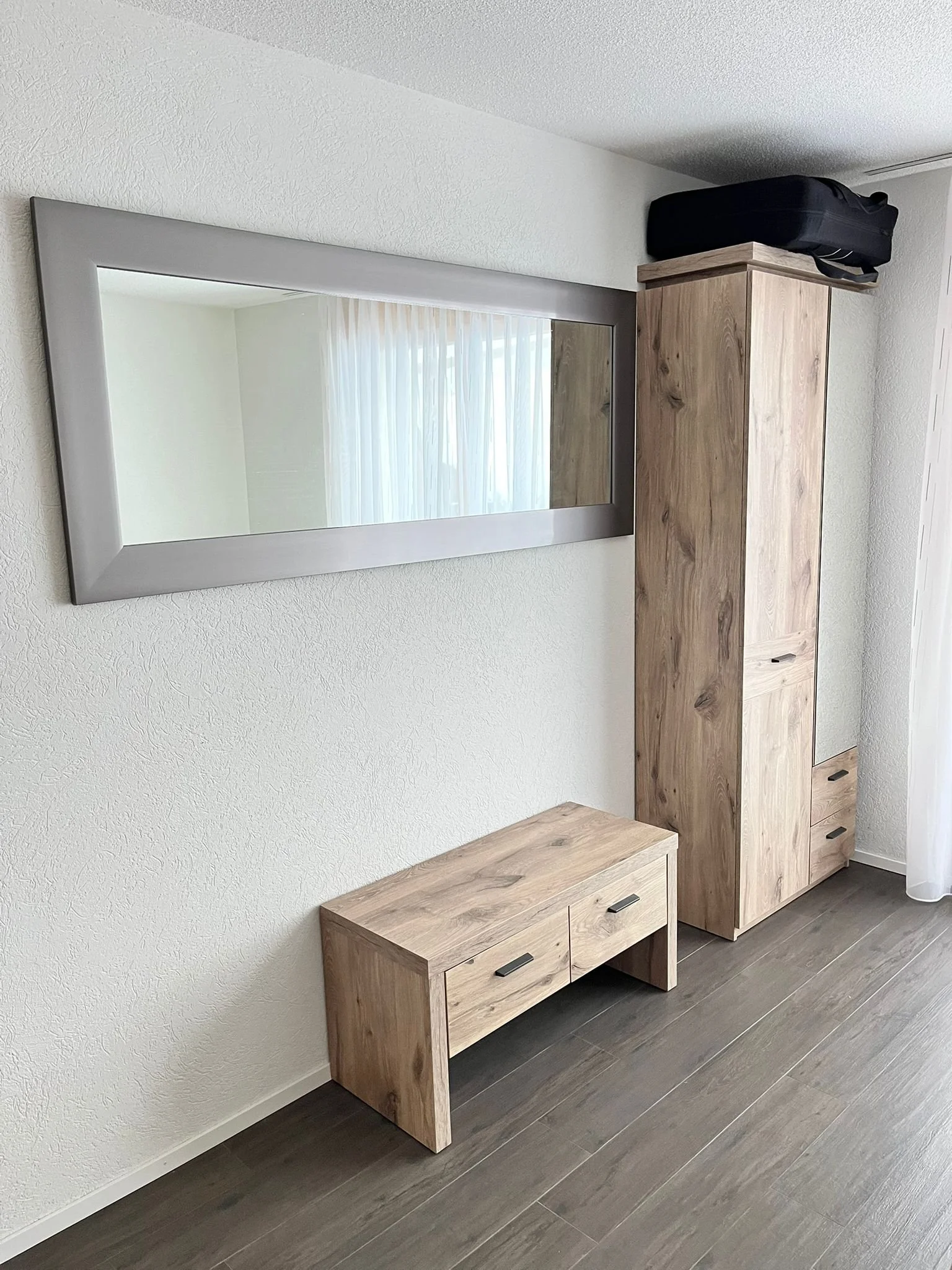 Ein grauer Wandspiegel hängt über einem kleinen Holztisch. Neben dem Tisch steht ein hoher Holzschrank und auf dem Schrank liegt ein schwarzer Koffer. Der Raum hat weiße Wände, grauen Holzboden und ist hell beleuchtet.