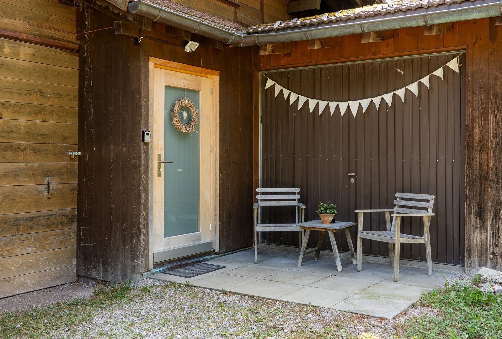 Ein gemütlicher Eingangsbereich mit Holzverkleidung, zwei Gartenstühlen, einem kleinen Holztisch mit Topfpflanze, einer Dekoration an der Tür und einer Wimpelkette.