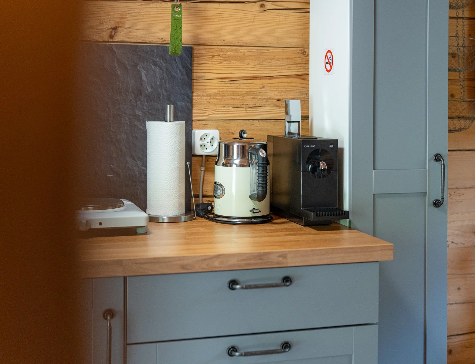 Küchenarbeitsfläche mit Kaffeepadmaschine, Wasserkocher, Papierhandtuchrolle und Steckdose, vor Holzwand und grauen Schränken.