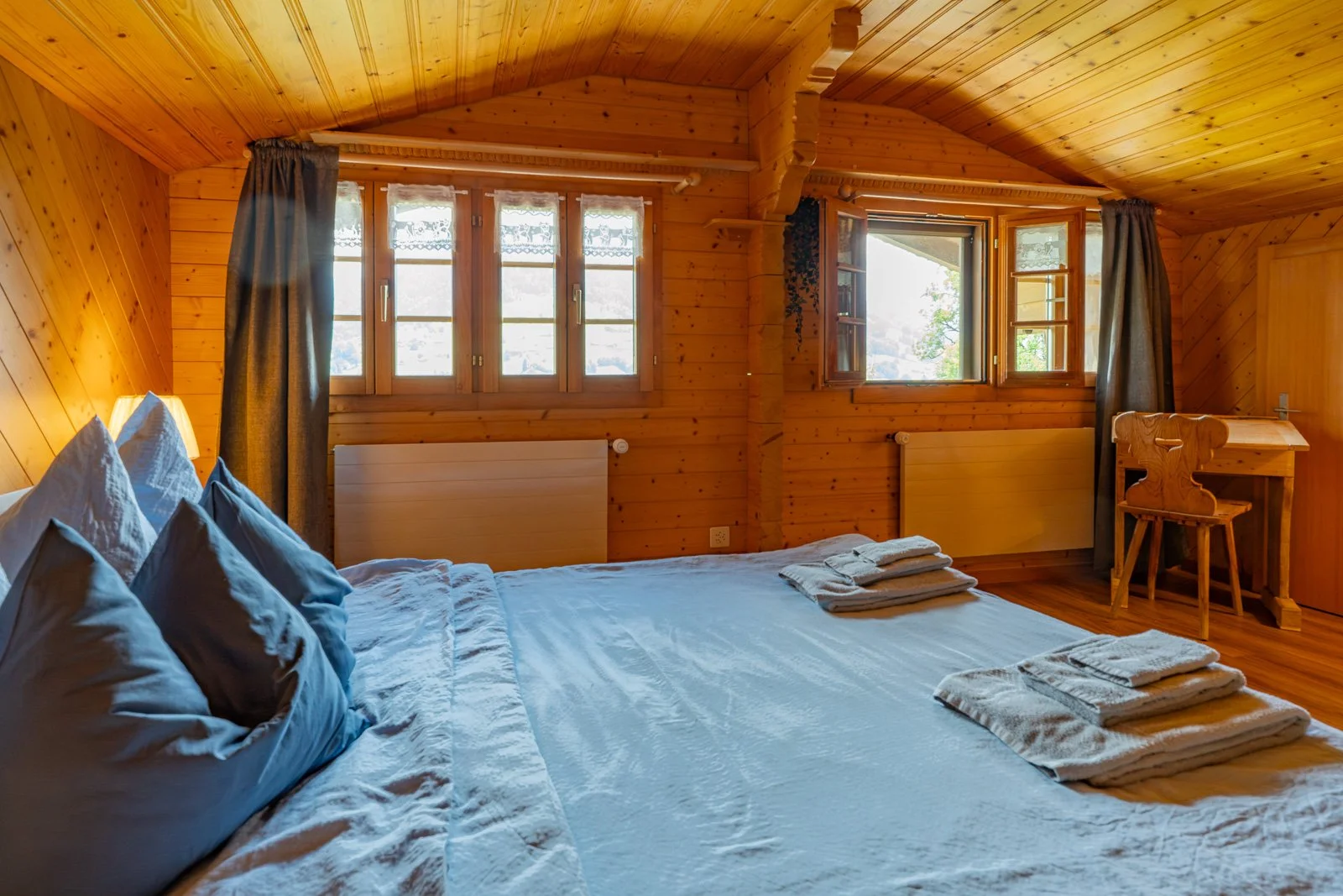 Ein Schlafzimmer mit Holzwänden, einem Bett mit weißen und blauen Kissen, mehreren handtuchartigen Gegenständen auf dem Bett, und drei Fenster, die Tageslicht hereinlassen, sowie ein kleiner Schreibtisch und Stuhl.