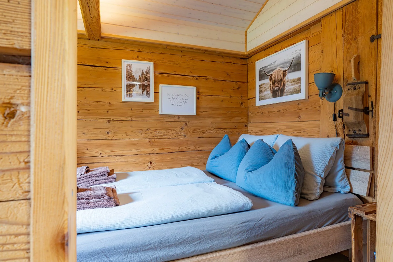 Ein Schlafzimmer mit Holzverkleidung, einem Bett mit weißen und blauen Kissen, eingerahmte Fotos an der Wand, darunter ein See, ein Zitat und eine Hochlandkuh.