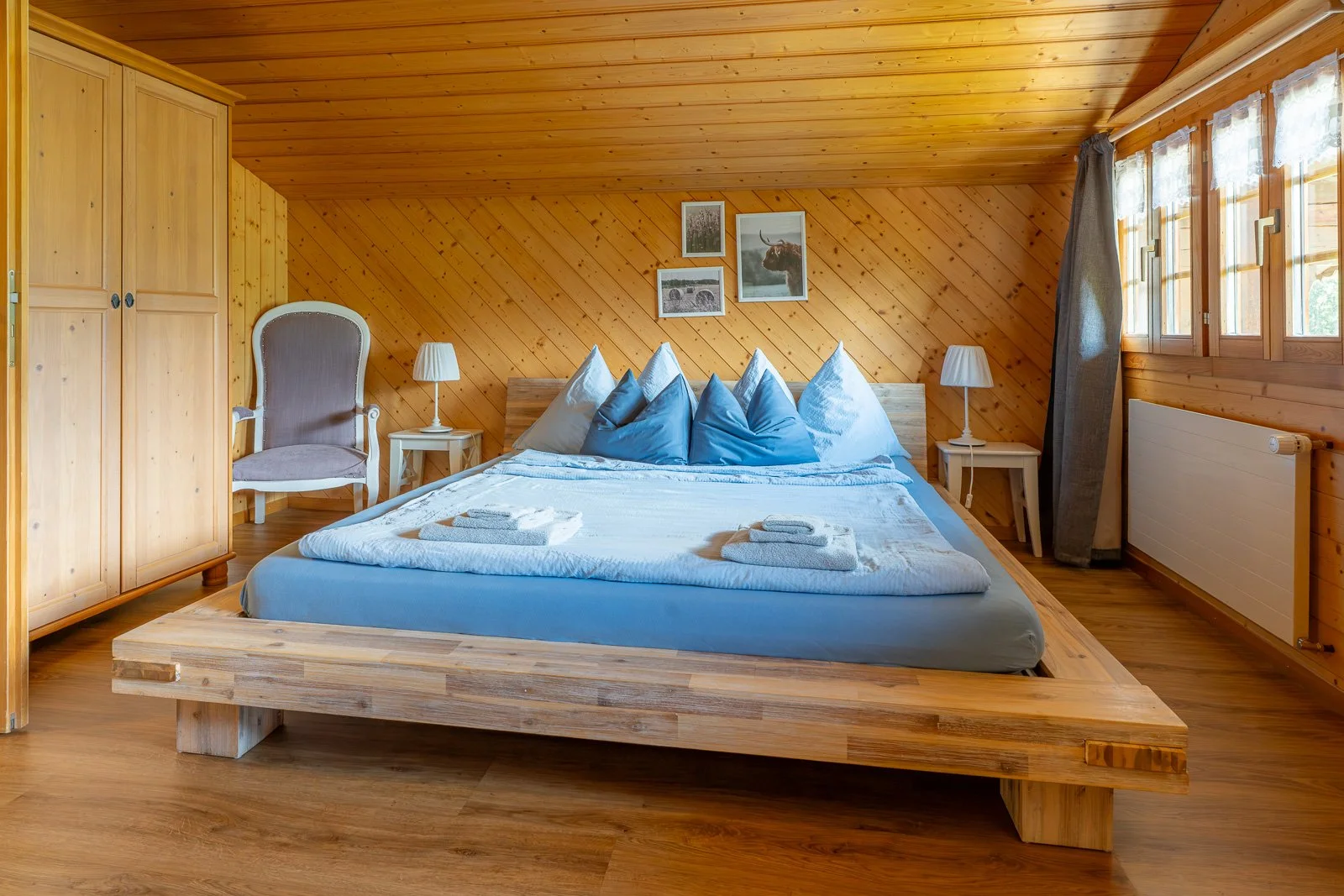 Ein gemütliches Schlafzimmer mit Holzvertäfelung, einem großen Bett mit blauen und weißen Kissen, Nachttischen mit Lampen, einem großen Kleiderschrank und Sonnenlicht, das durch die Fenster scheint.