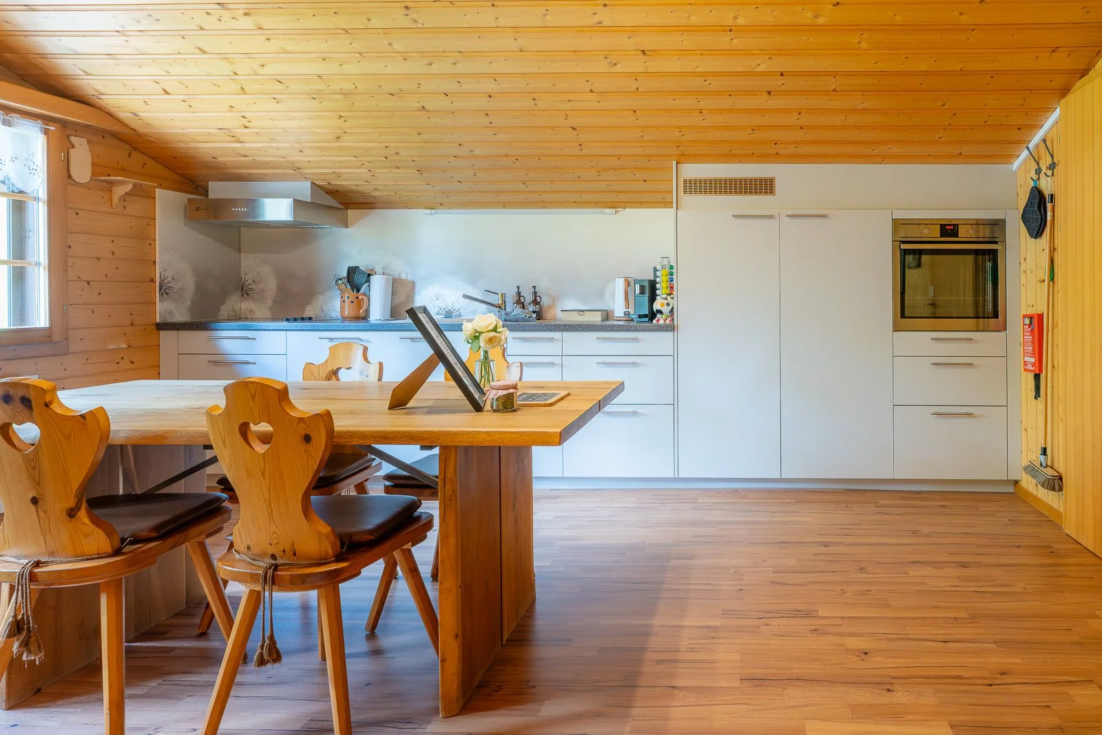Innenraum einer modernen Küche mit Holzdecke, Holzfußboden und weißem Küchenmobiliar. Es gibt einen Esstisch aus Holz mit Stühlen, und die Küche ist mit modernen Geräten und Dekorationen ausgestattet.