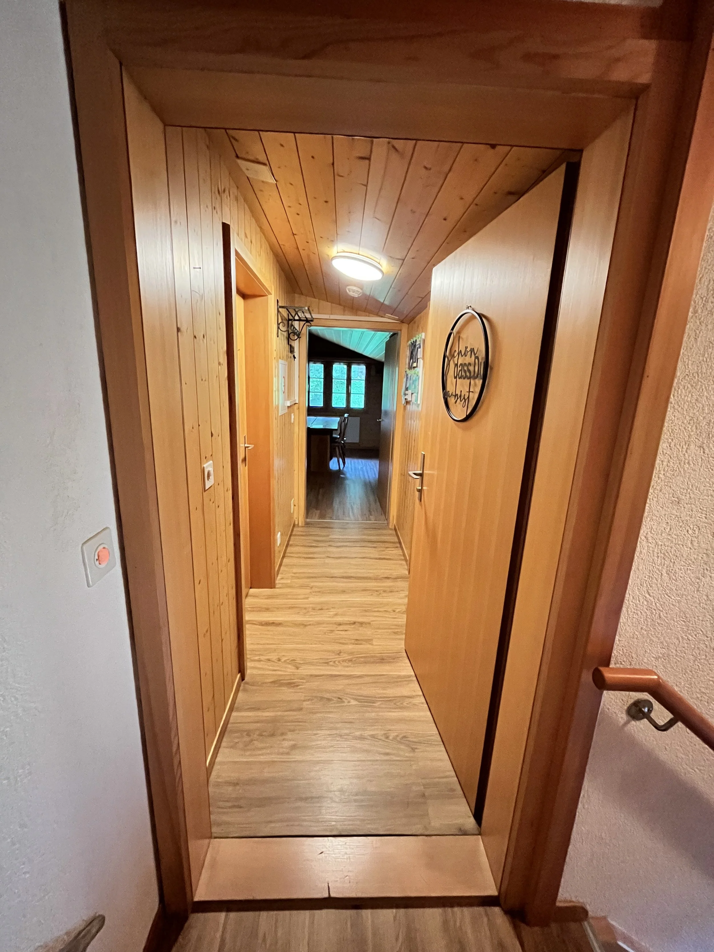 Holzverkleideter Flur in einem Haus mit Blick auf einen Raum mit großen Fenstern im Hintergrund.