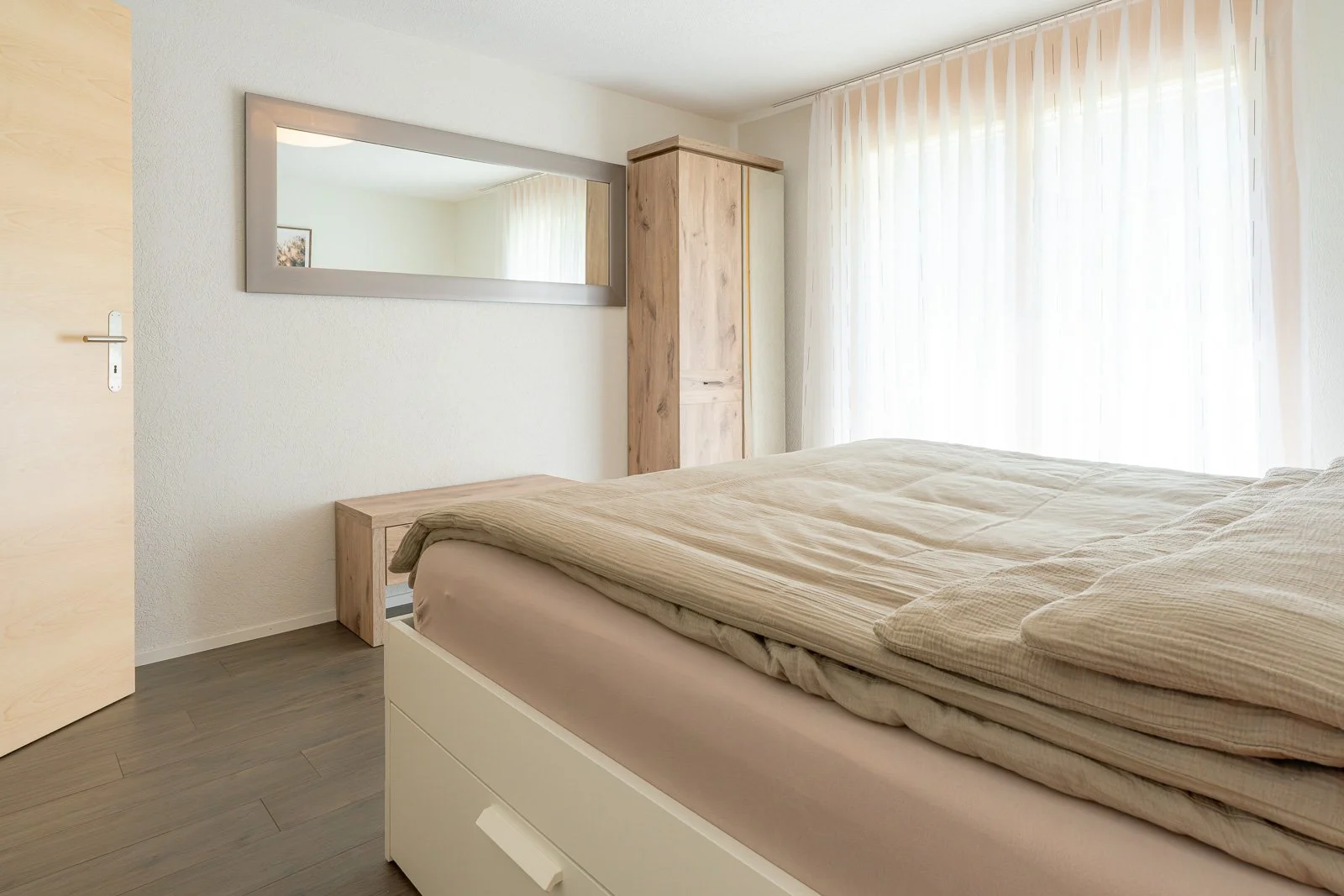 Schlafzimmer mit Bett, Wandspiegel, Holzmöbeln und großen Fenstern mit weißen Vorhängen