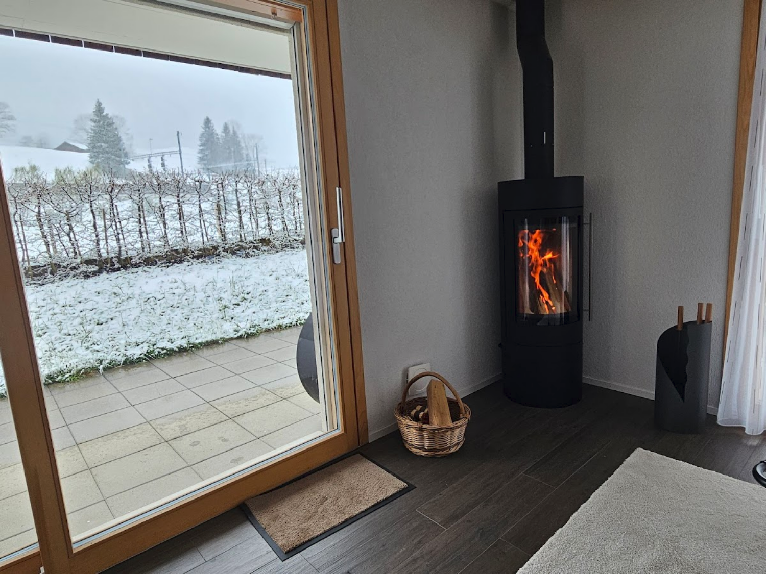 Innenraum mit einem Holzofen, der Feuer hat, neben einer kleinen Korbtasche mit Holzscheiten, vor einem Fenster mit Blick auf Schnee und Bäume im Winter.