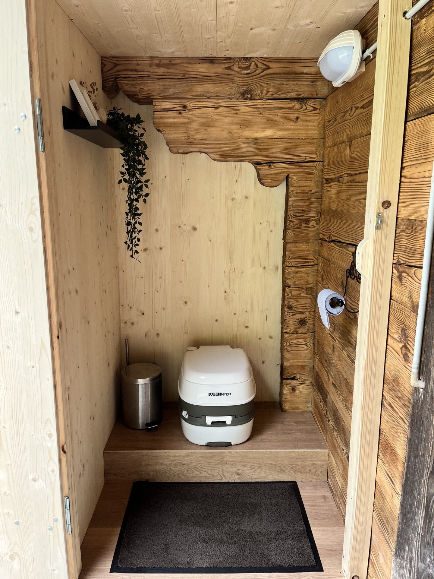 Ein kleines Toilettenhäuschen mit Holzvertäfelung, darin eine moderne tragbare Toilette, einen Mülleimer, eine Toilettepapierrolle und eine kleine Ablage mit Pflanzen.
