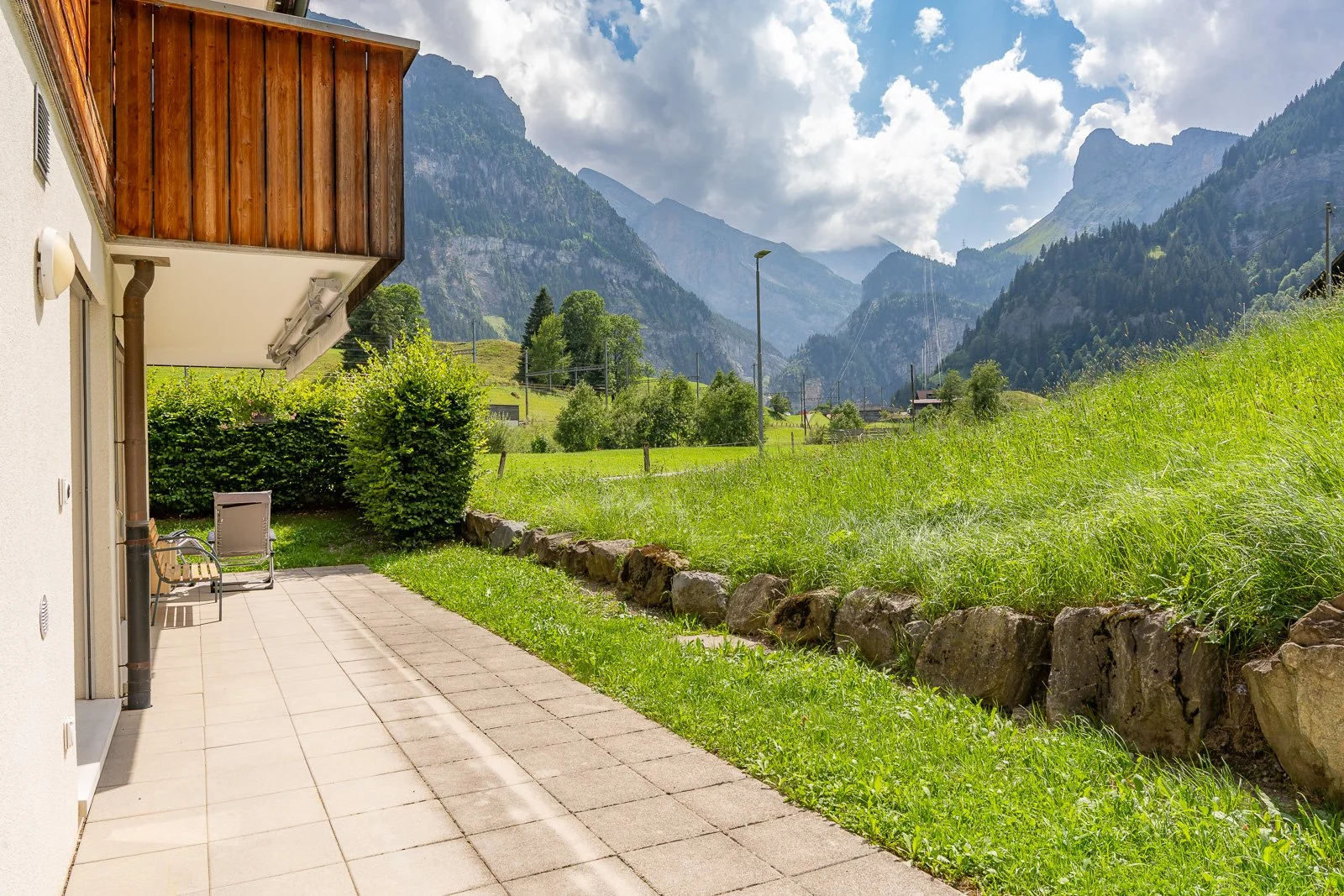 Balkon mit Blick auf eine grüne Wiese und Berge im Hintergrund, sonniger Tag, einige Bäume und Wolken am Himmel.