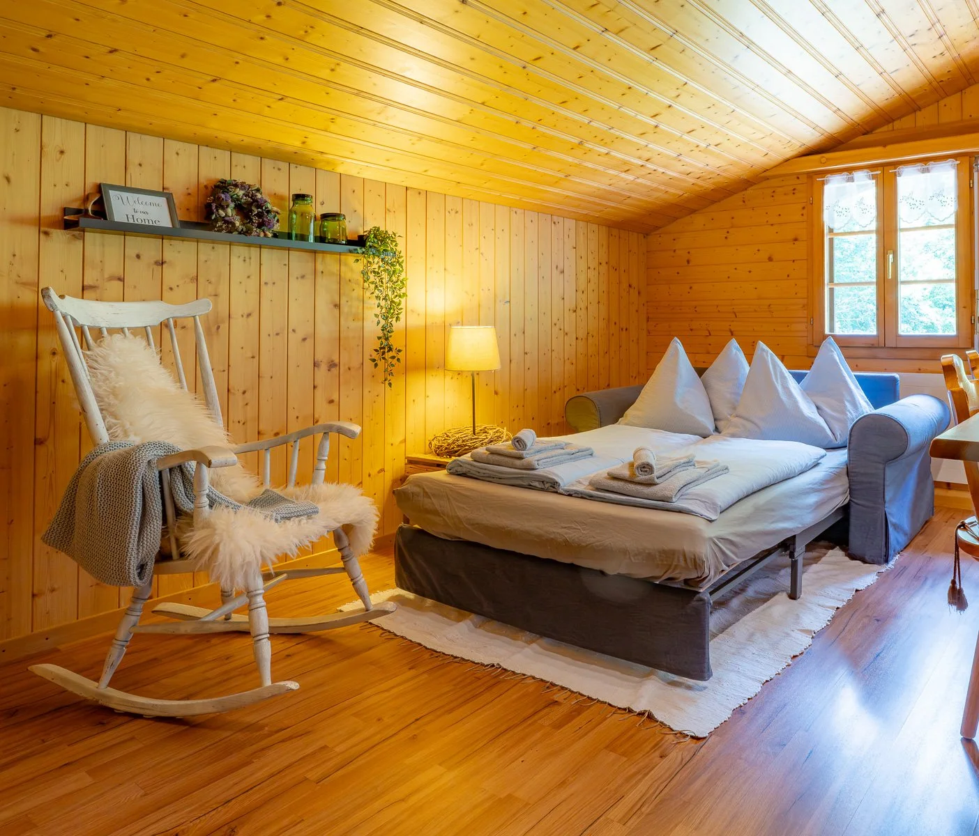 Gemütliches Schlafzimmer in einem Holzhaus mit einem Doppelbett, einem Sessel mit Fellbezug, einem Regal mit Dekorationen, einem Nachttisch mit Lampe, einem Fenster und Holzvertäfelung an den Wänden und Decke.