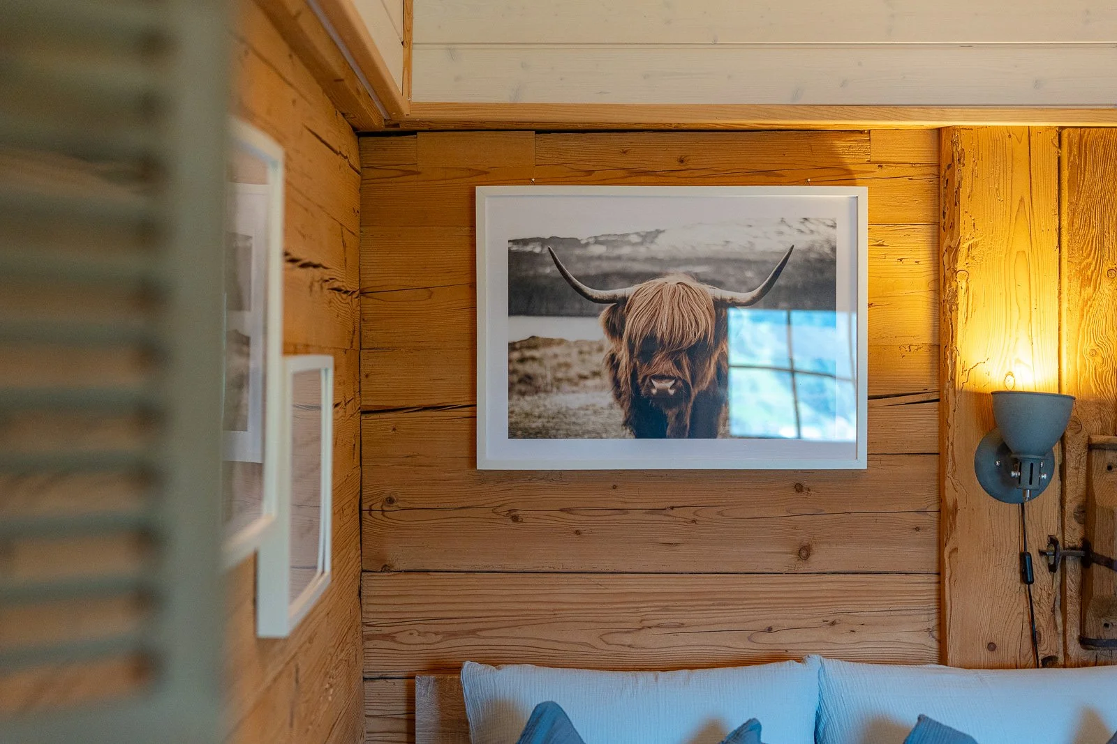 Ein Wandbild eines Scotland Highland Rinderkopfes an einer Holzwand in einem gemütlichen Raum, auf der rechts ein Wandlampenarm mit Leuchte sichtbar ist.