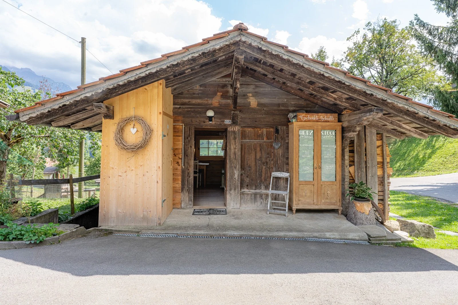 Ein Holzhaus mit einem Dach, dekoriert mit einem Herz aus Zweigen an der linken Seite und einem Holzschrank mit Schildern auf der rechten Seite, umgeben von Bäumen und Pflanzen, mit einem Himmel im Hintergrund.