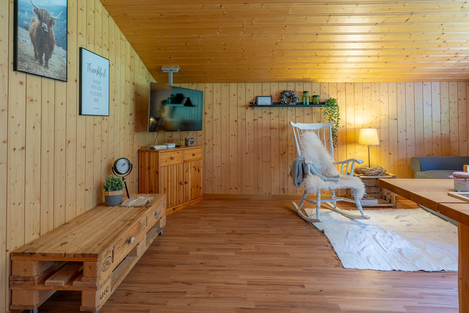 Innenaufnahme eines gemütlichen Wohnzimmers mit Holzwänden, einer Schaukelstuhl mit Kissen, einem Fernseher, einem Bücherregal, einer Stehlampe, einem Sofa und einem Couchtisch.