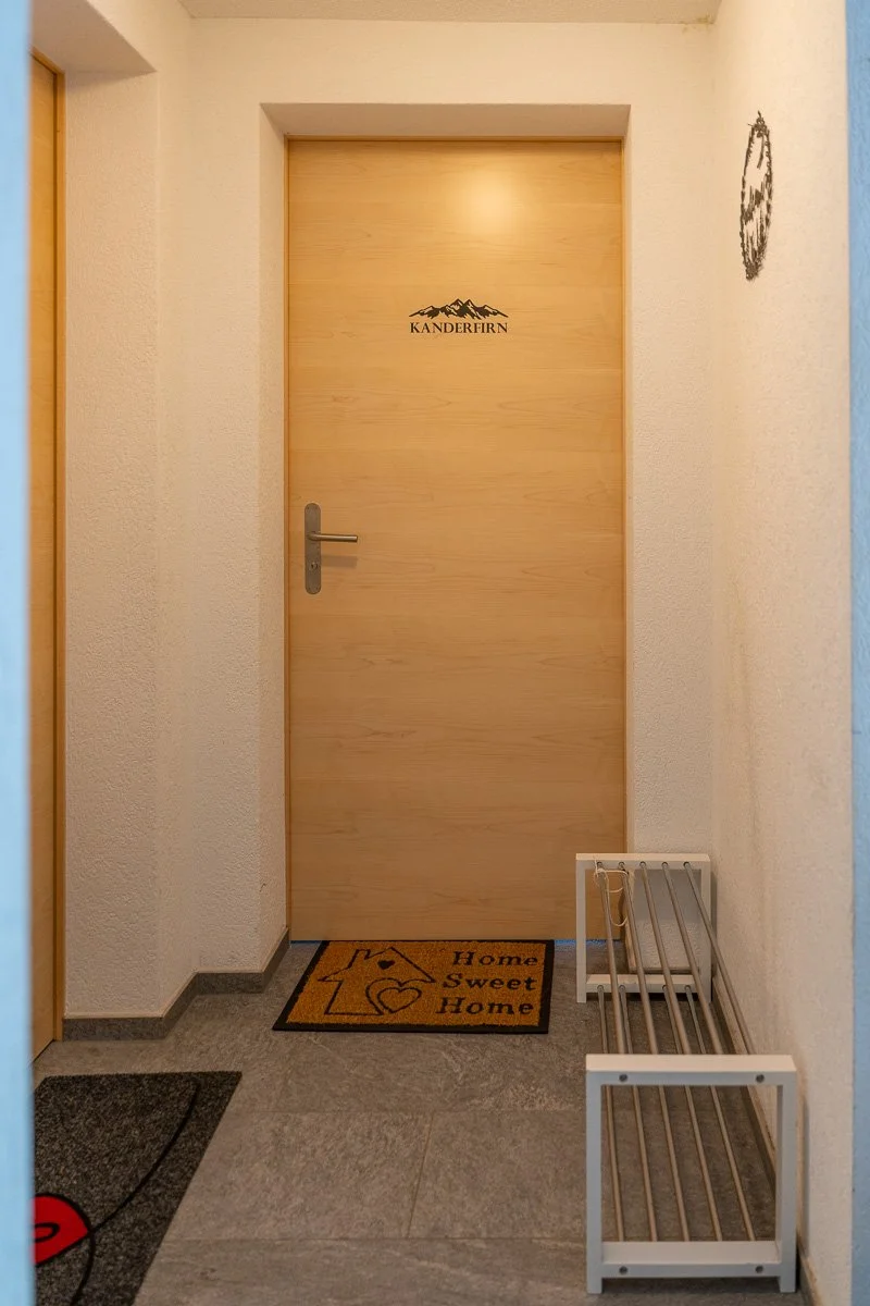 Holztür mit dem Logo "KANDERFIRN" und einem Wandbild auf der rechten Seite, davor ein Begrüßungsmattentuch mit der Aufschrift "Home Sweet Home". Der Eingangsbereich ist schlicht eingerichtet mit einem Schuhregal auf der rechten Seite.
