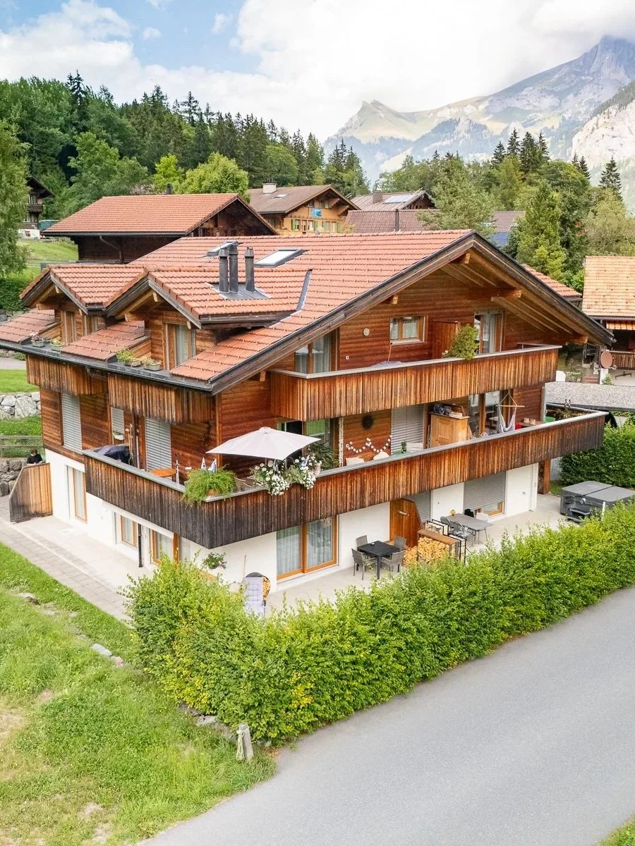 Ein Holzhaus mit Dachterrasse und Balkon vor einer grünen Landschaft mit Bäumen und Bergen im Hintergrund.