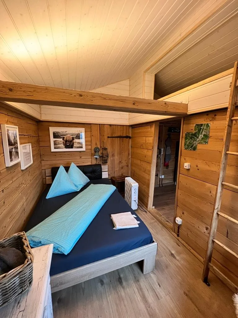 Ein Holzwohnzimmer mit einem Doppelbett, dekoriert mit blauen Kissen und Bettwäsche, an einer Wand hängen Fotos, und eine Leiter führt zu einem oberen Bereich.