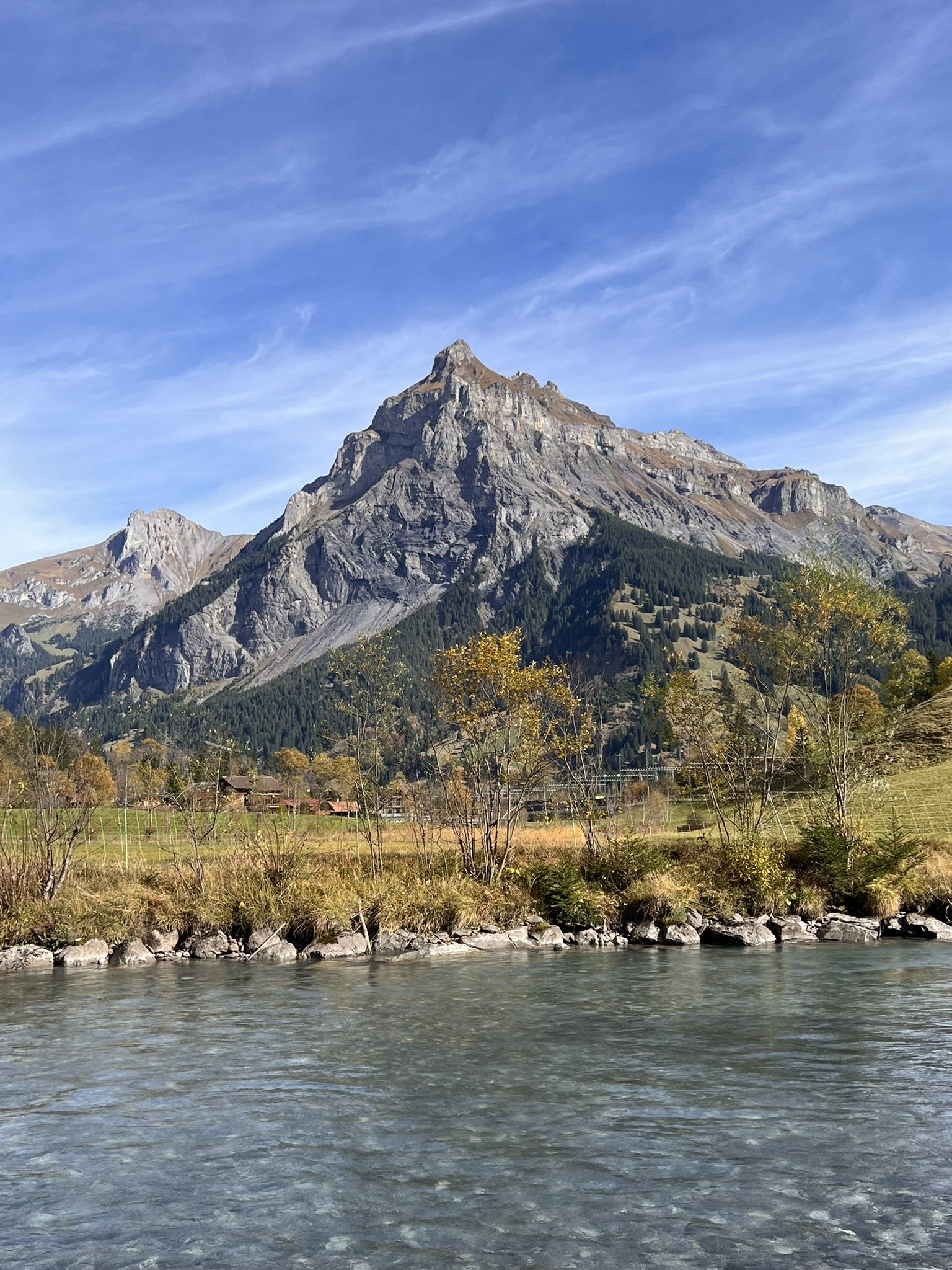 Berg mit steilen Felswänden, umgeben von Wäldern und einer Wiese, im Vordergrund ein Fluss, blauer Himmel mit wenigen Wolken.