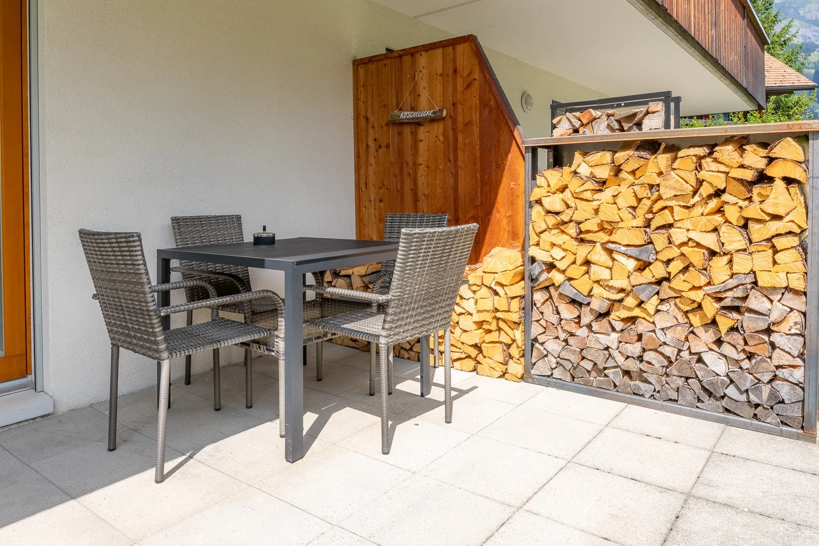 Blick auf einen Balkon mit einem schwarzen Esstisch, vier Stühlen aus Rattan, einer Holzscheitaufbewahrung, einem Holzregal mit Holzscheiten, und einem Holzschild mit der Aufschrift Kaschtecke.