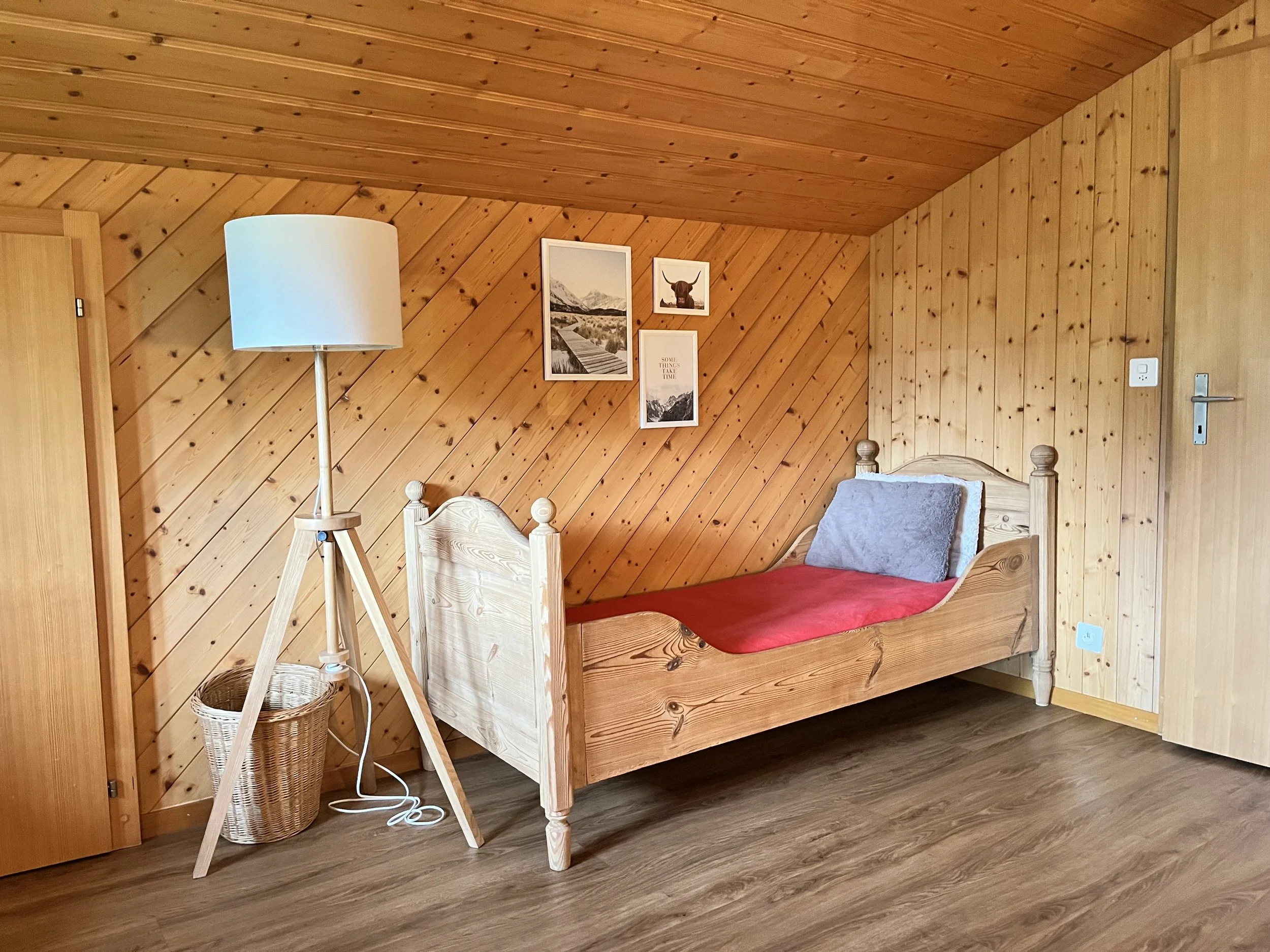 Ein Schlafzimmer mit Holzverkleidung an den Wänden und der Decke. Es gibt ein Einzelbett mit einer grauen und einer weißen Decke. Neben dem Bett steht eine Stehlampe mit einem weißen Lampenschirm. Auf der Wand hängen vier Bilder, darunter ein Bergbil
