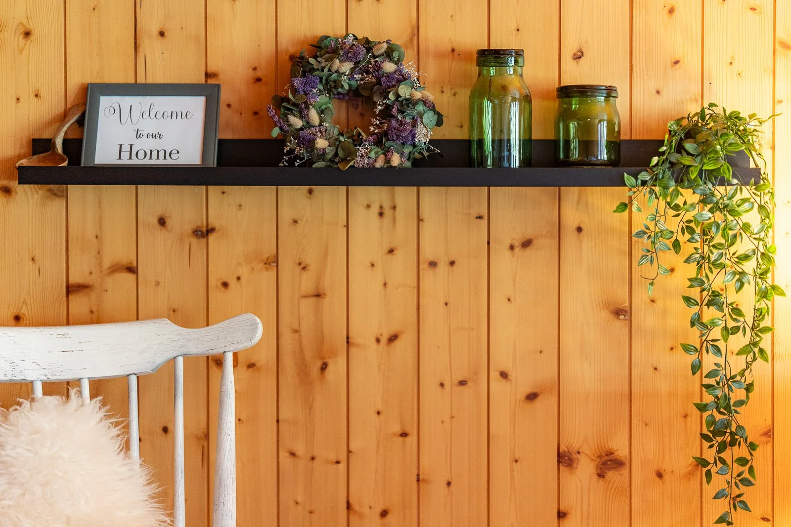 Eine Holzregal mit einem Willkommensschild, einem Blumenkranz, zwei grünen Gläsern und einem kleinen Topfpflanze. Im Vordergrund ein weißer Stuhl mit Fell. Die Wand ist aus Holzbrettern.