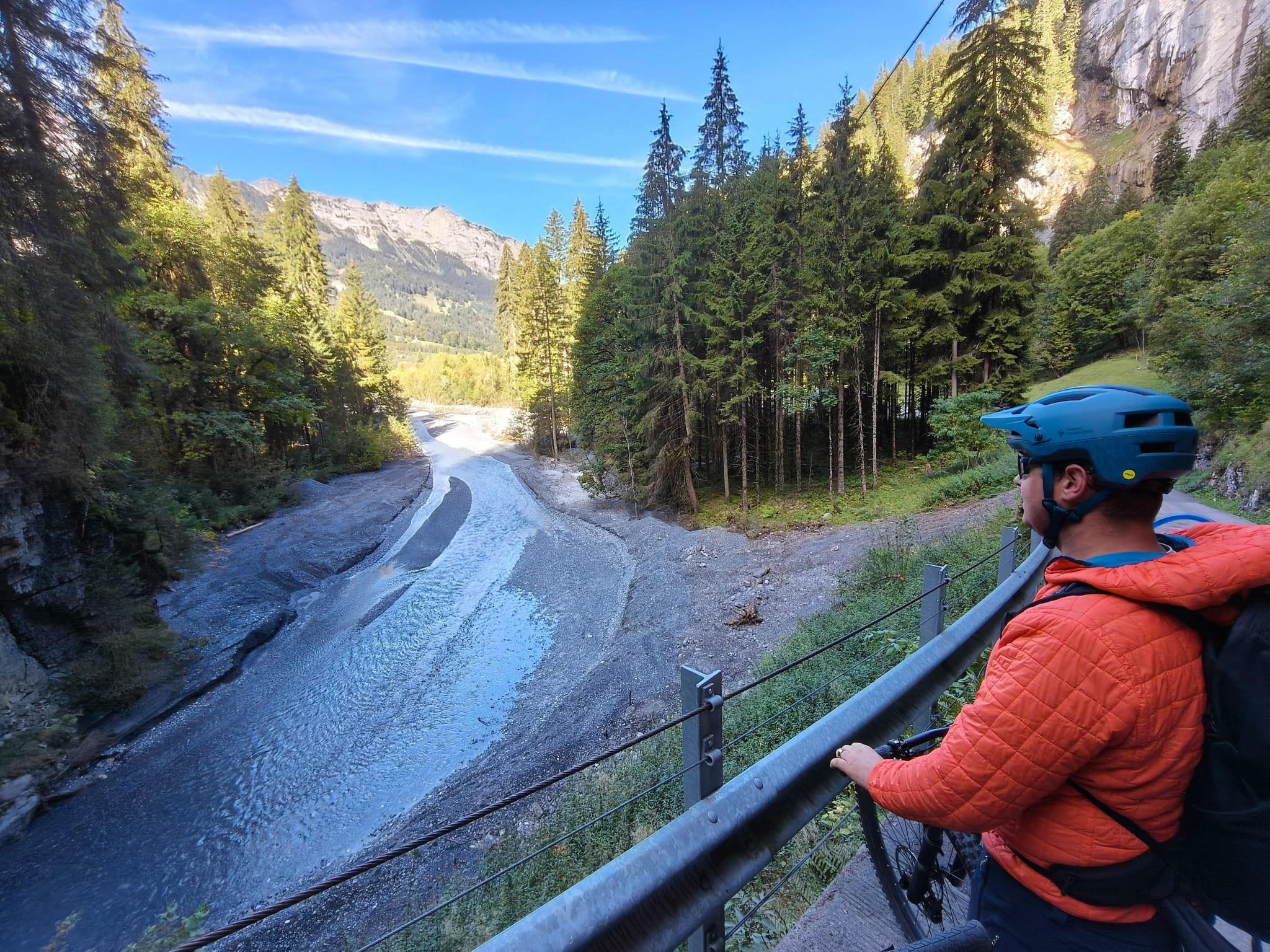Mountainbiker mit blauem Helm und orangefarbener Jacke an einer Schlucht mit Wildbach, Nadelwald und alpiner Felslandschaft