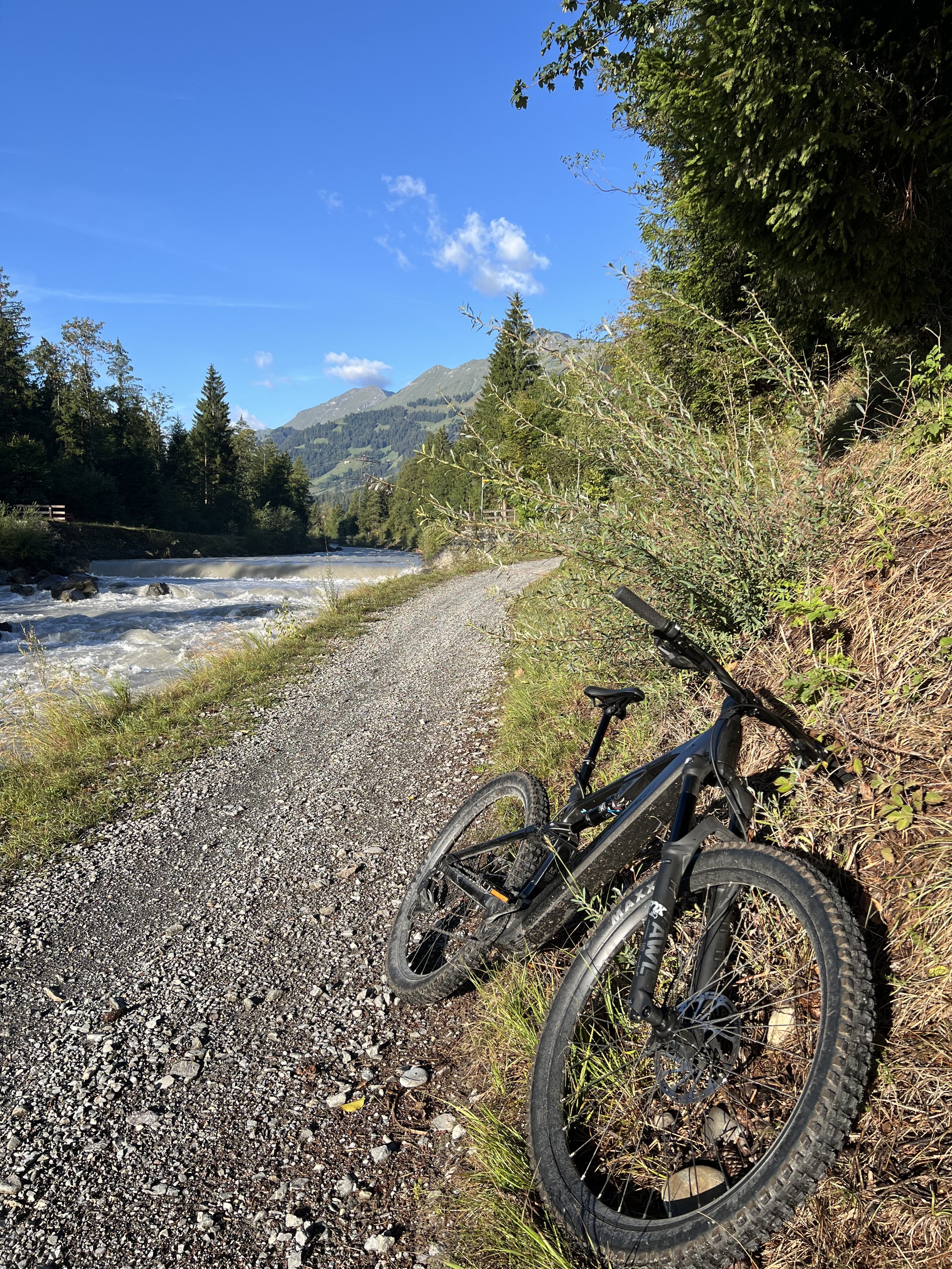 Mountainbike auf einem Schotterweg am Fluss vor bewaldeten Alpen und blauem Himmel
