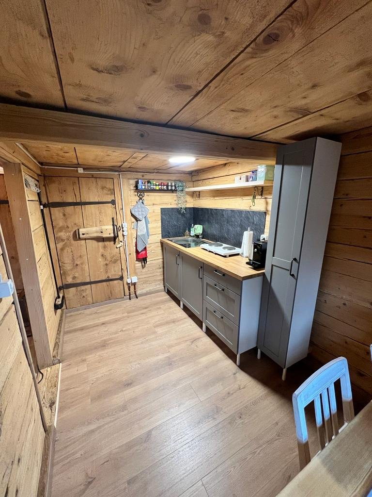 Küche mit Holzwänden und -decken, grauem Küchenmöbel, kleiner Arbeitsfläche, Backofen, Kaffeemaschine, Geschirrspüler, offene Regale mit Vorräten, aufgeräumt, gemütlich.