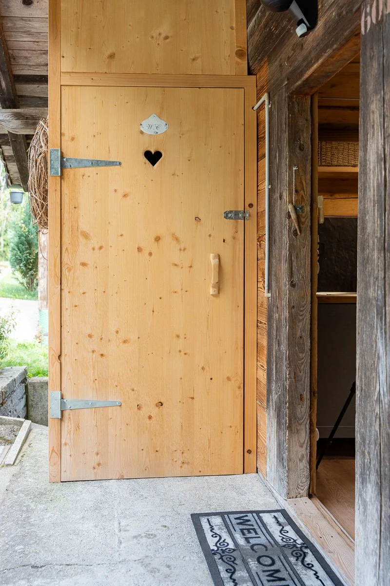 Eine Holztür mit einem kleinen Herzfenster und einem Willkommensmatte davor, die Tür befindet sich an einer Holzveranda.