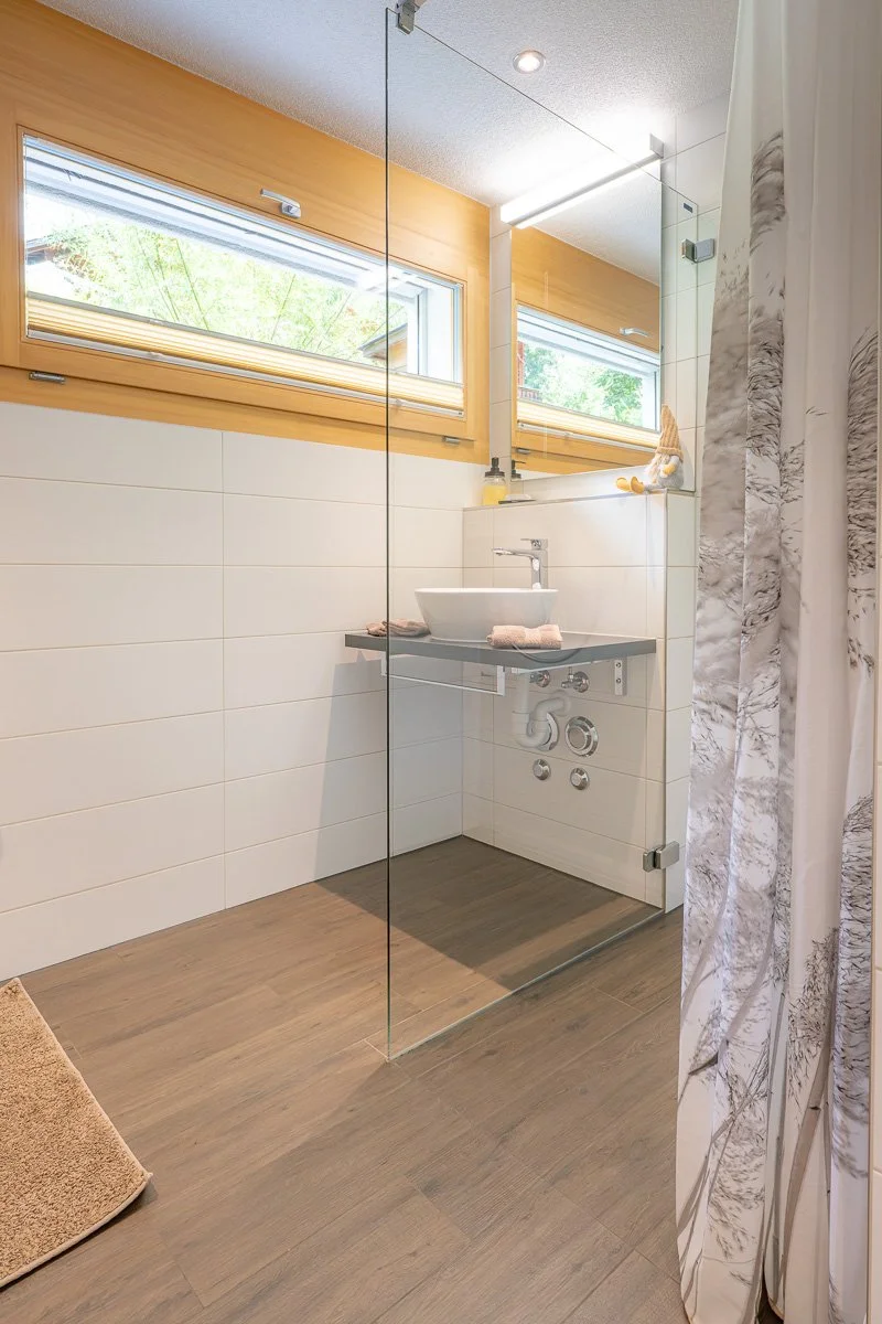 Moderne Dusche mit Glaswand, Holzfußboden, weißer Wandfliese, Fenster mit Holzrahmen, Waschbecken auf schwarzem Regal, Spiegel, Duschvorhang mit Marmoroptik.
