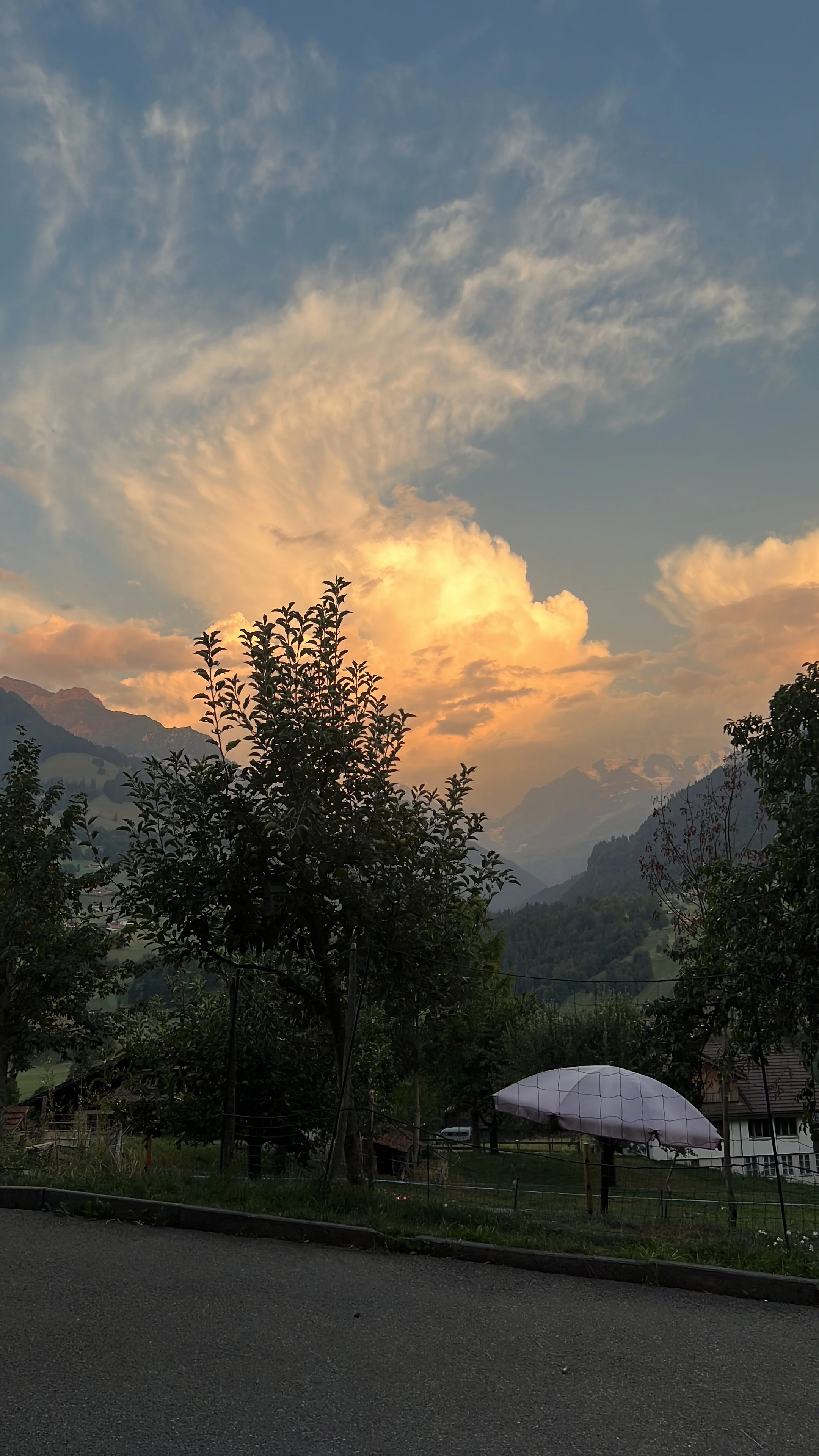 Sonnenuntergang mit leuchtenden Wolken über einer Berglandschaft, Obstbäumen, Gewächshaus und ländlichen Häusern