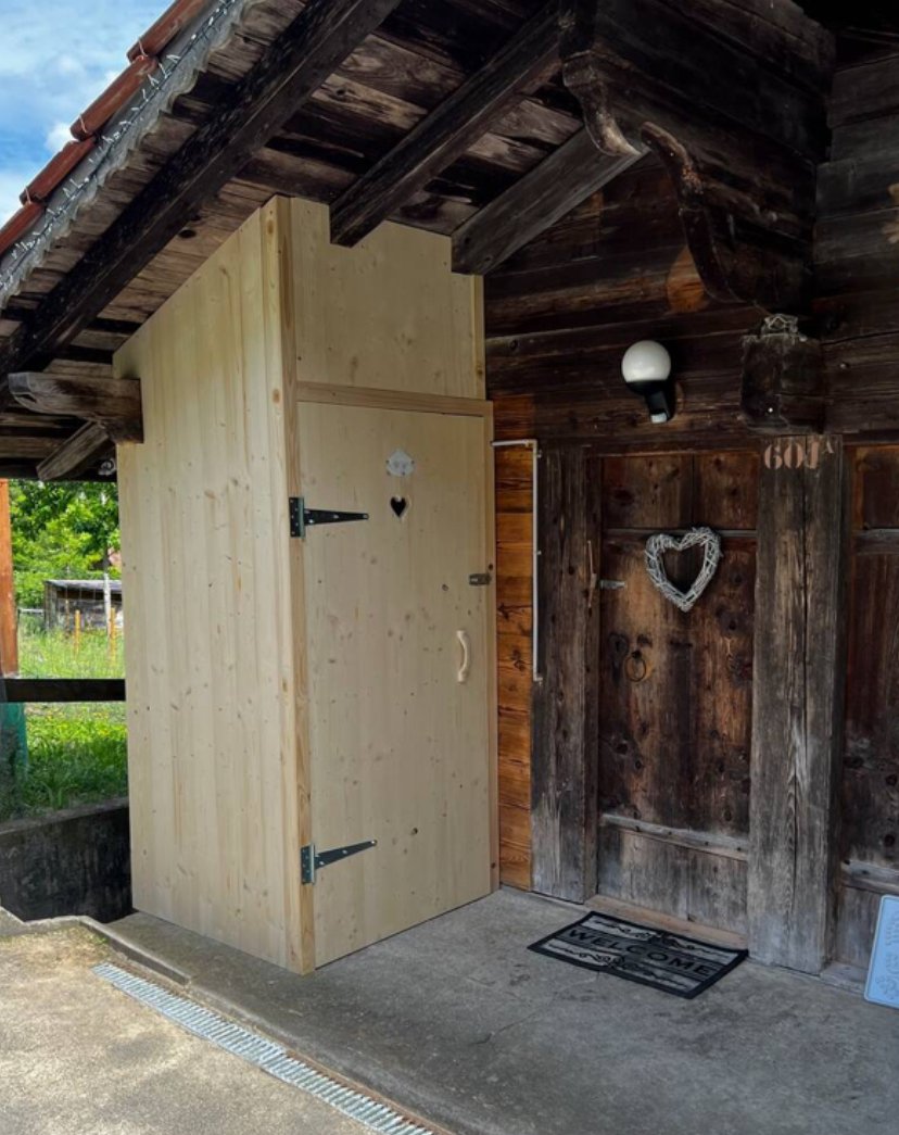 Gartendusche mit Holzverkleidung und einer transparenten Tür, unter einem Dach aus dunklem, gebrauchten Holz, mit einem Begrüßungsmatten davor.