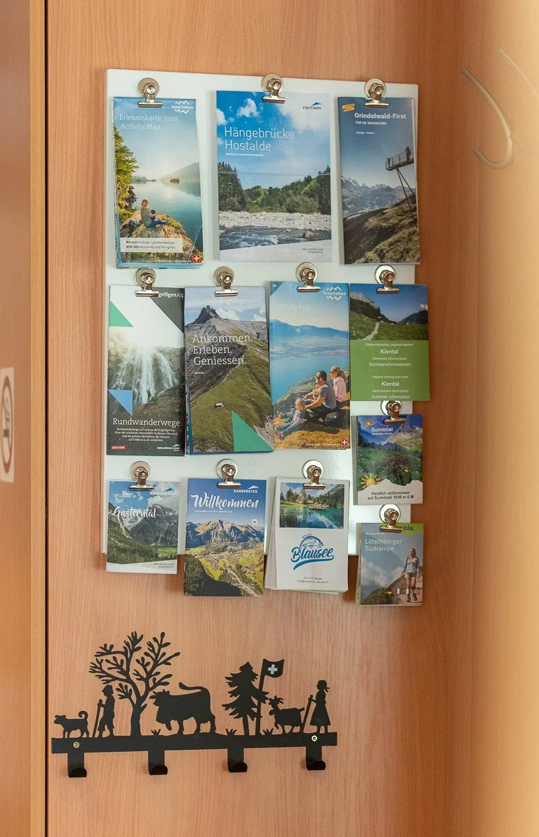 Informationsbroschüren und Karten für Touristen in einer Holztafel, darunter eine Holzschnitt-Deko mit Schweizer Motiven, wie Bär, Hirsch und Bergsteiger, auf einer Wand.