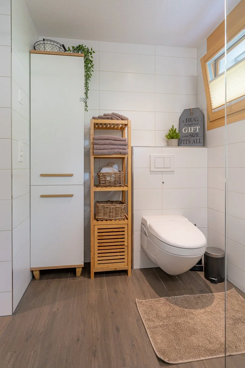 Ein Badezimmer mit weißen Fliesen, einer Toilette, einem Holzregal mit Handtüchern und Körben, einer kleinen Pflanze und einem Wandbild mit dem Zitat "A hug is a great gift, one size fits all". Es gibt eine kleine Fenster mit Holzrahmen und einen bra