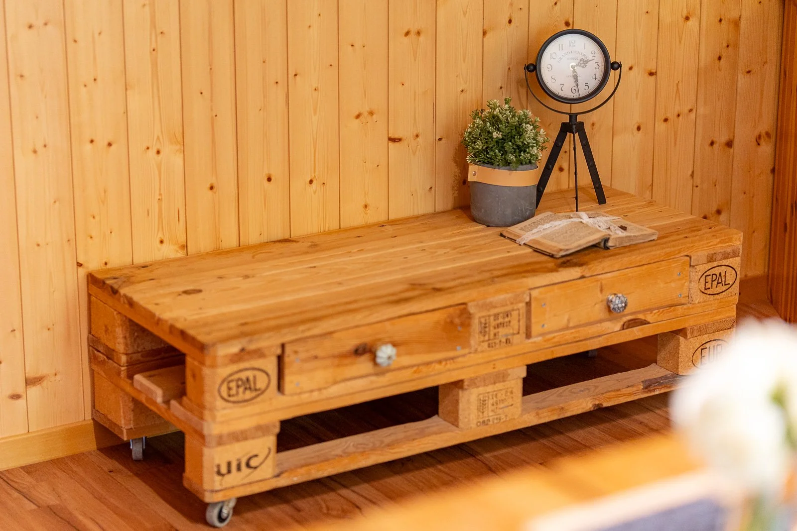 Ein Holztisch aus Weinkisten mit Schubladen, auf dem eine Pflanze, eine alte Uhr und ein offenes Buch liegen, vor einer Holzvertäfelung.