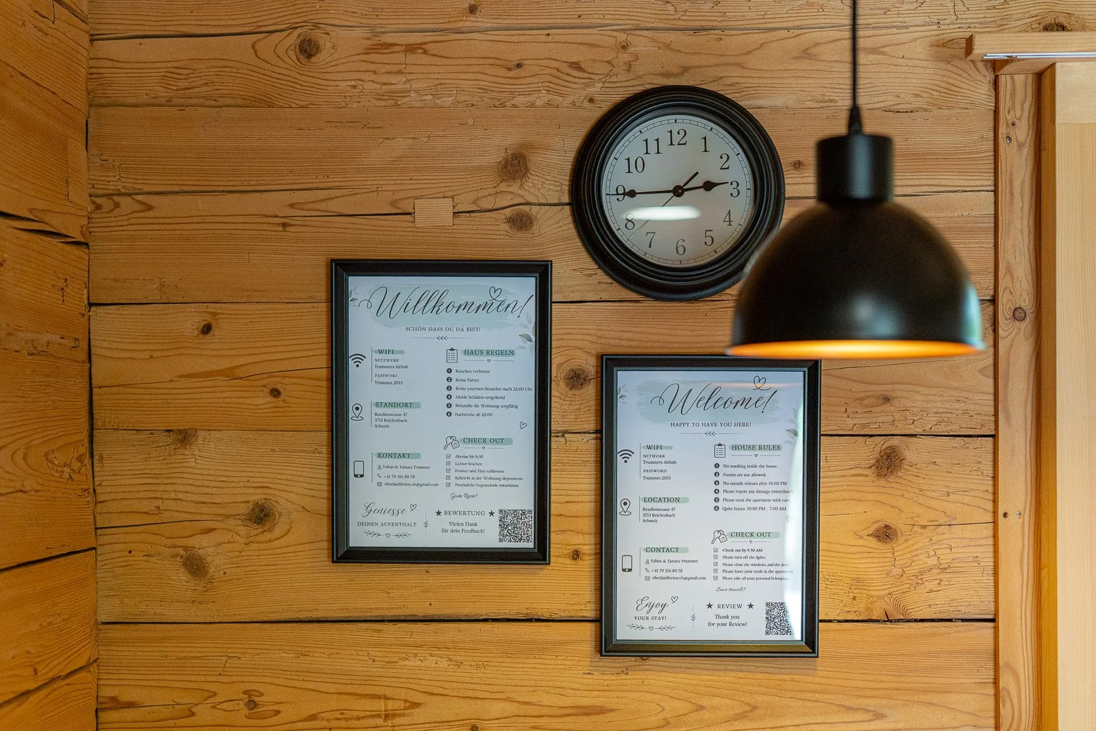 Holzwand mit zwei schwarzen Bildern mit deutschen und englischen Texten, einer Uhr und einer schwarzen Lampe. Die Bilder enthalten Willkommens- und Verhaltensregeln sowie Kontaktinformationen.