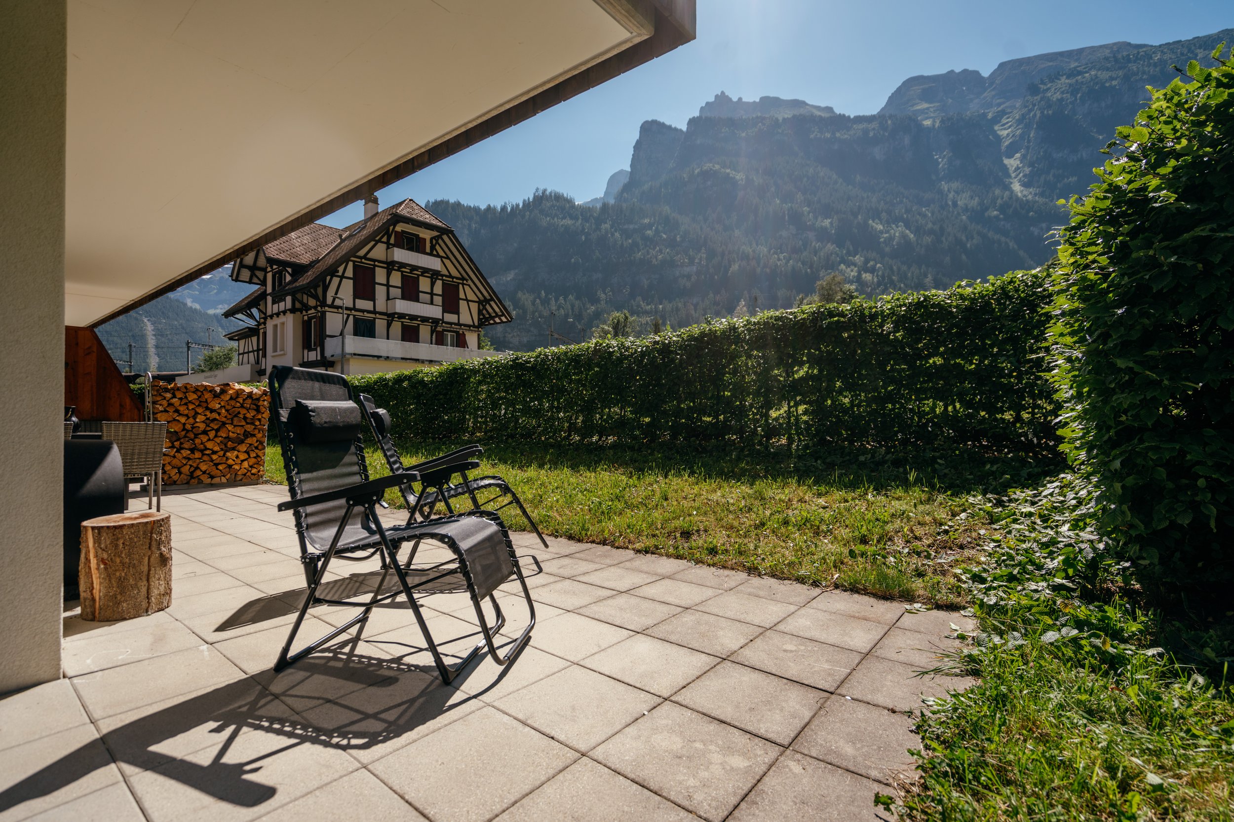 Blick von einer Terrasse auf ein traditionelles Chalethaus mit Holzofenstapel, umgeben von einer grünen Hecke, mit Bergen im Hintergrund, bei sonnigem Wetter.