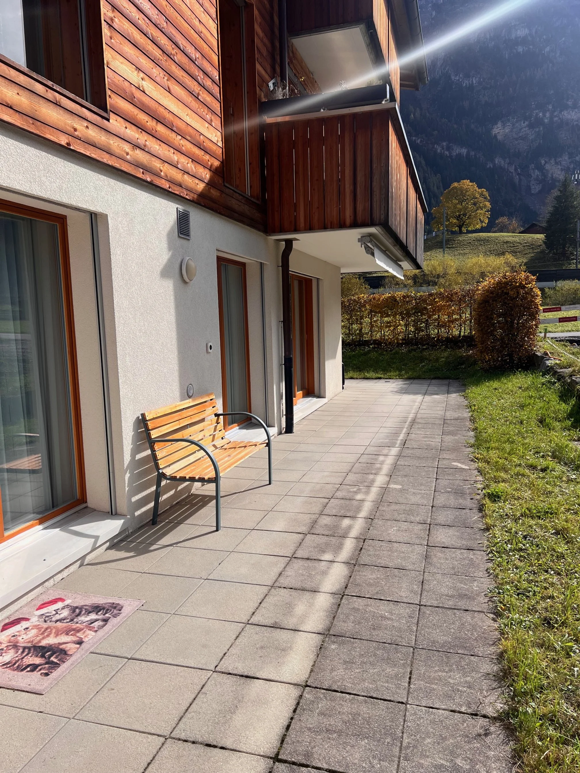Balkon mit Holzmöbel vor einem modernen Haus mit Holz und Putz an der Fassade, umgeben von grünen Büschen und Bergen im Hintergrund, sonniger Tag.