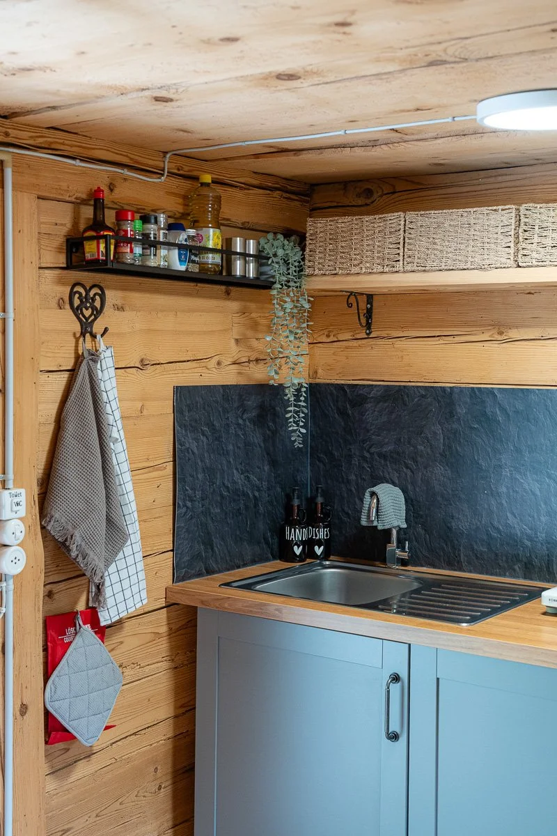 Küchenecke mit Holzverkleidung, Spüle, blaue Unterschränke, schwarzen Wandplatte, Kräutertöpfen, Geschirrtüchern, einem Regal mit Gewürzen und sonstigen Küchenutensilien, einer Lampe an der Decke.