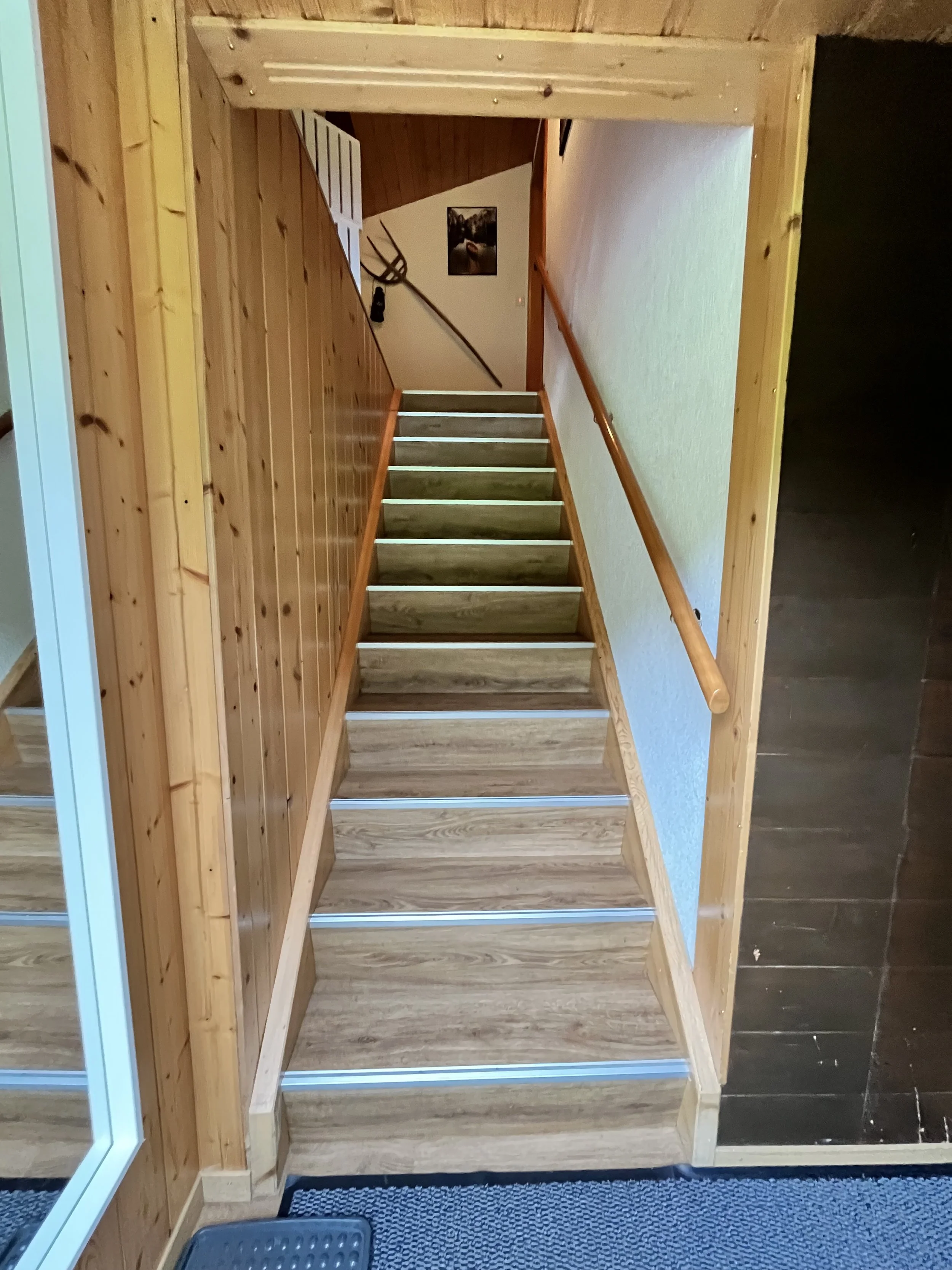 Holztreppe mit Metallverstärkungen, Holzhandlauf und Holzverkleidung an den Wänden, Blick nach oben, auf der Wand oben befindet sich eine Wanddekoration mit einer Sense und einer Schaufel.