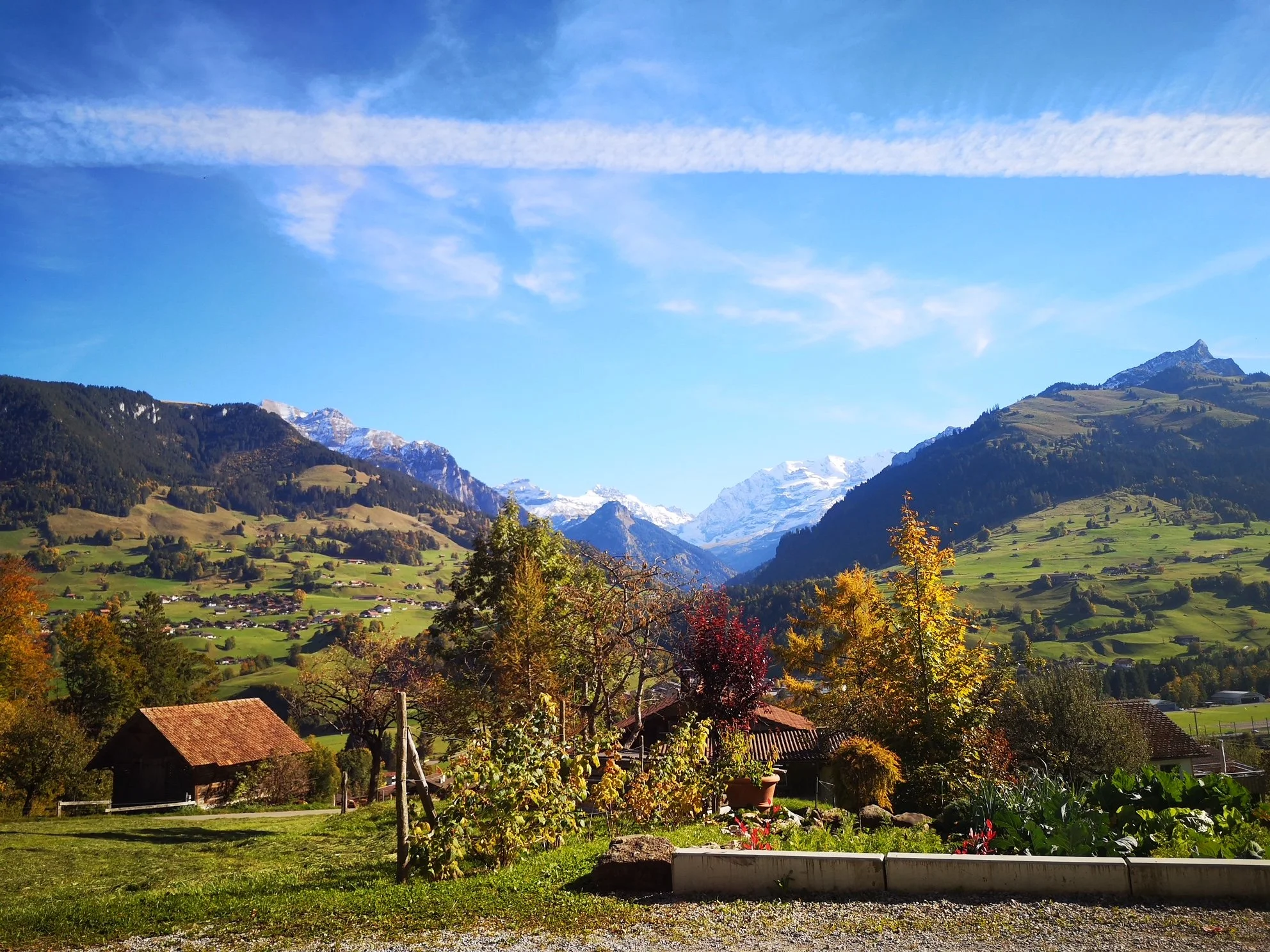 Berglandschap mit grünen Hängen, bunten Bäumen, Häusern im Vordergrund und schneebedeckten Bergen im Hintergrund bei sonnigem Himmel.