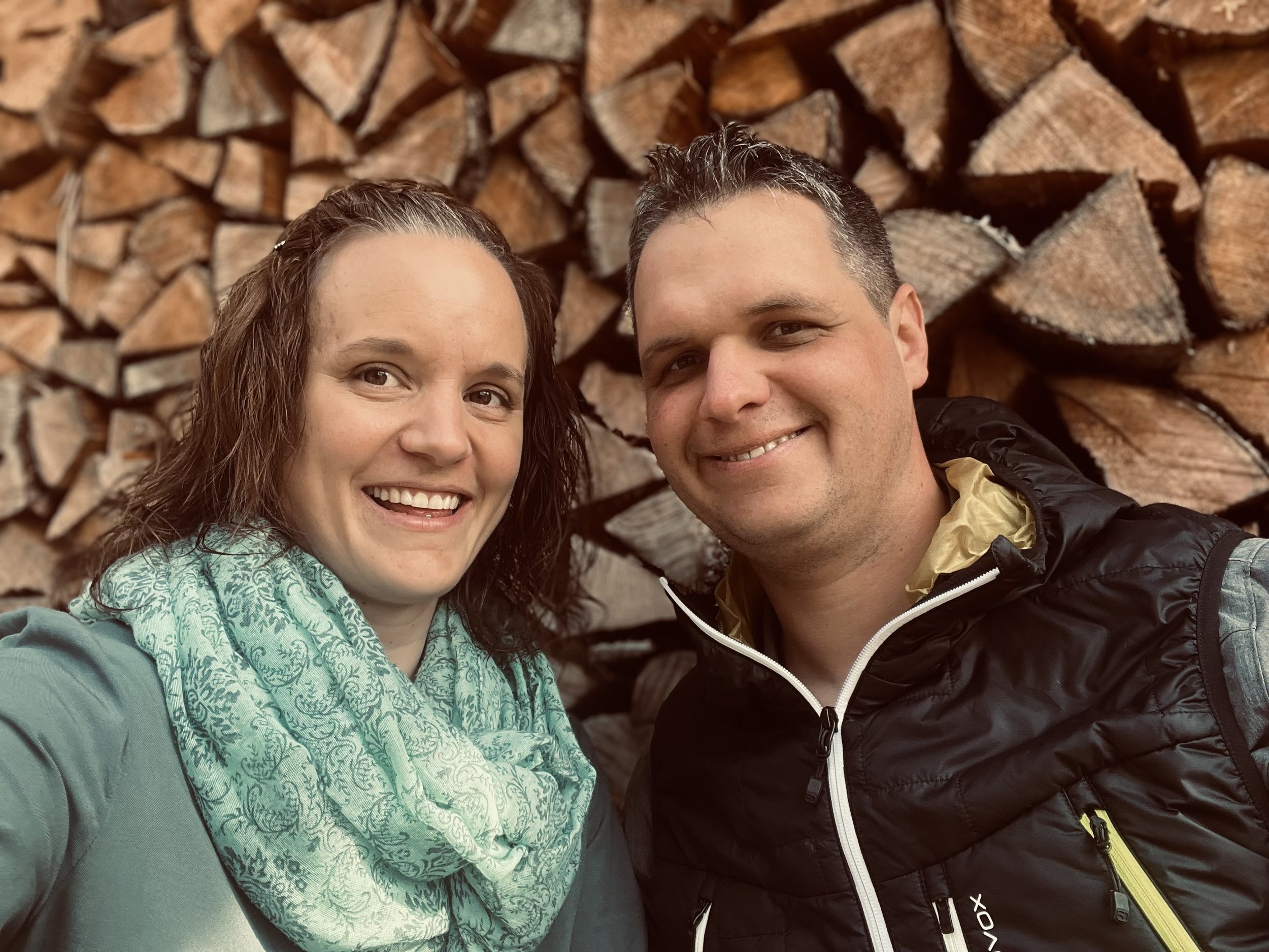 Ein lachendes Paar vor einem Stapel Holzscheite, beide sind mittleren Alters und tragen outdoor Kleidung.