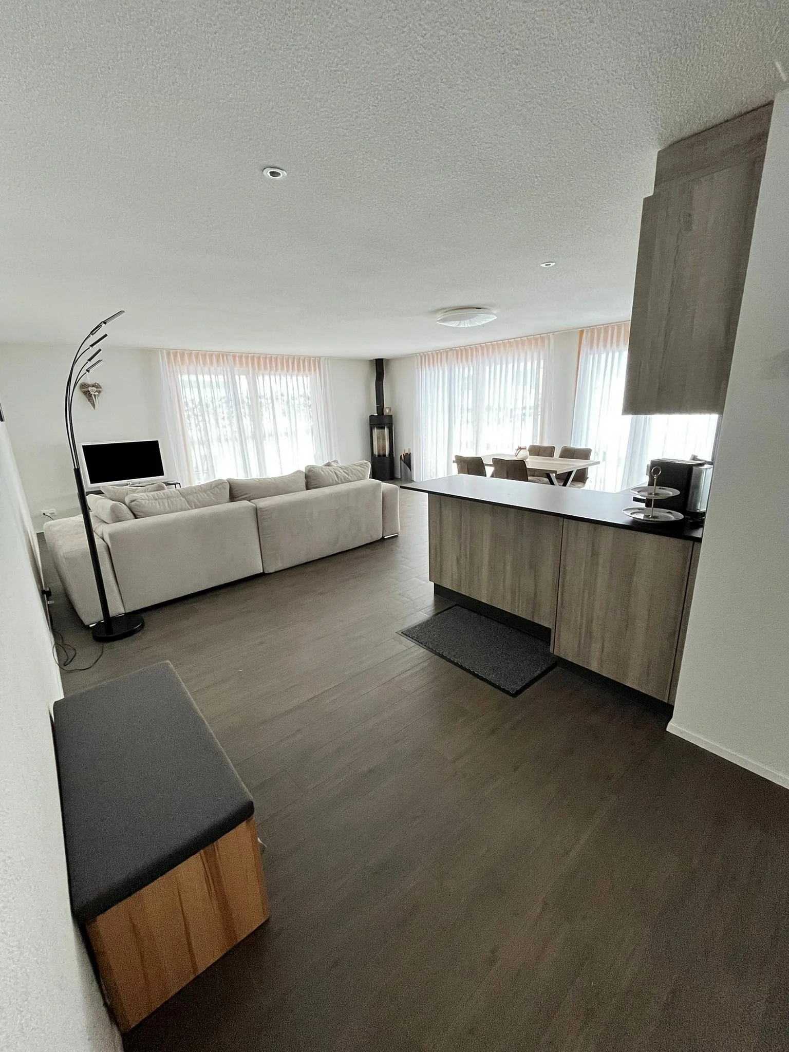 Ein heller, moderner Wohnraum mit einem weißen Sofa, einem Fernseher, einem Kamin, einem Esstisch mit Stühlen und großen Fenstern mit Vorhängen, die viel Tageslicht hereinlassen.