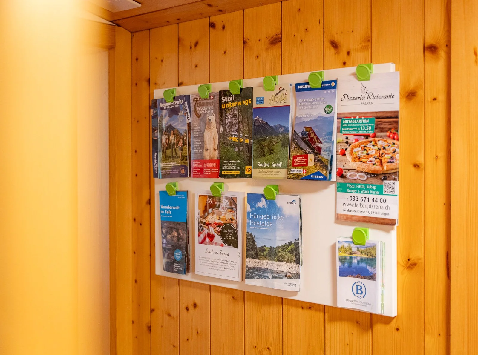 Ein Holzbrett an einer Wand mit verschiedenen Flyern und Broschüren, die Ausflugsziele, Pizzaspezialitäten und touristische Informationen zeigen.